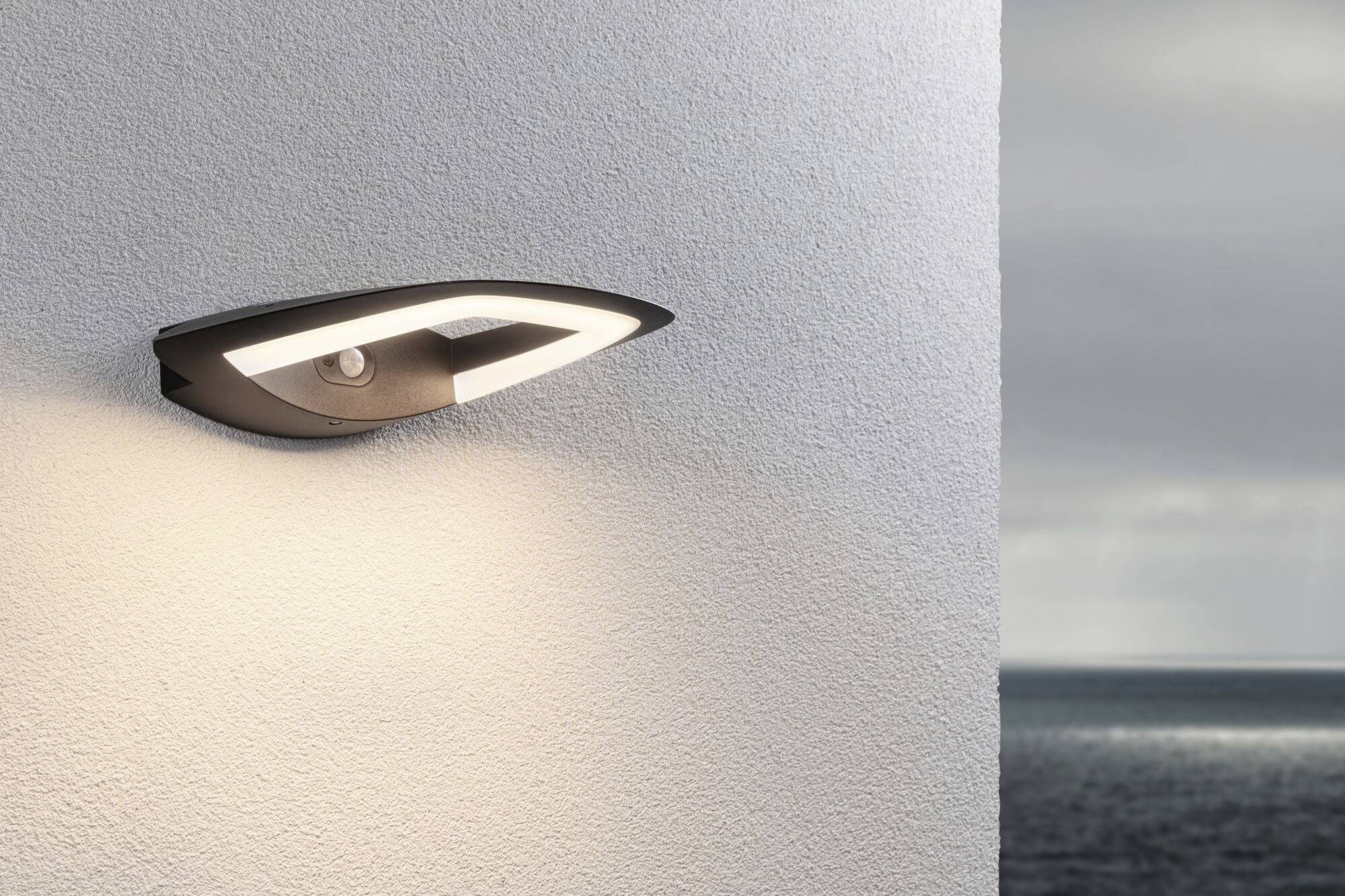 Een moderne buitenlamp aan een witte muur verlicht subtiel de omgeving. Op de achtergrond is de zee zichtbaar.