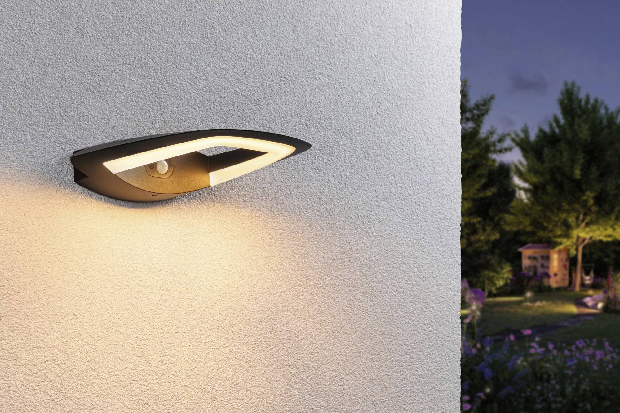 Een moderne buitenwandlamp verlicht een gevel in het schemerdonker. Op de achtergrond zijn bomen en een kleine tuinschuur te zien.