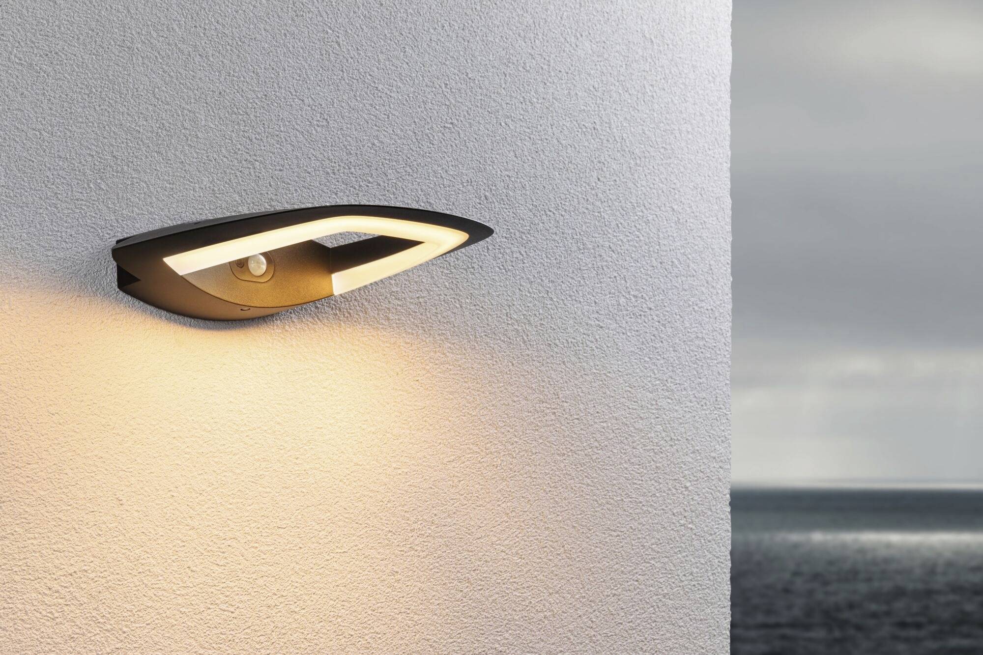 Moderne buitenwandlamp aan een witte muur, verlicht zacht het gebied voor een kustlandschap met een bewolkte lucht op de achtergrond.