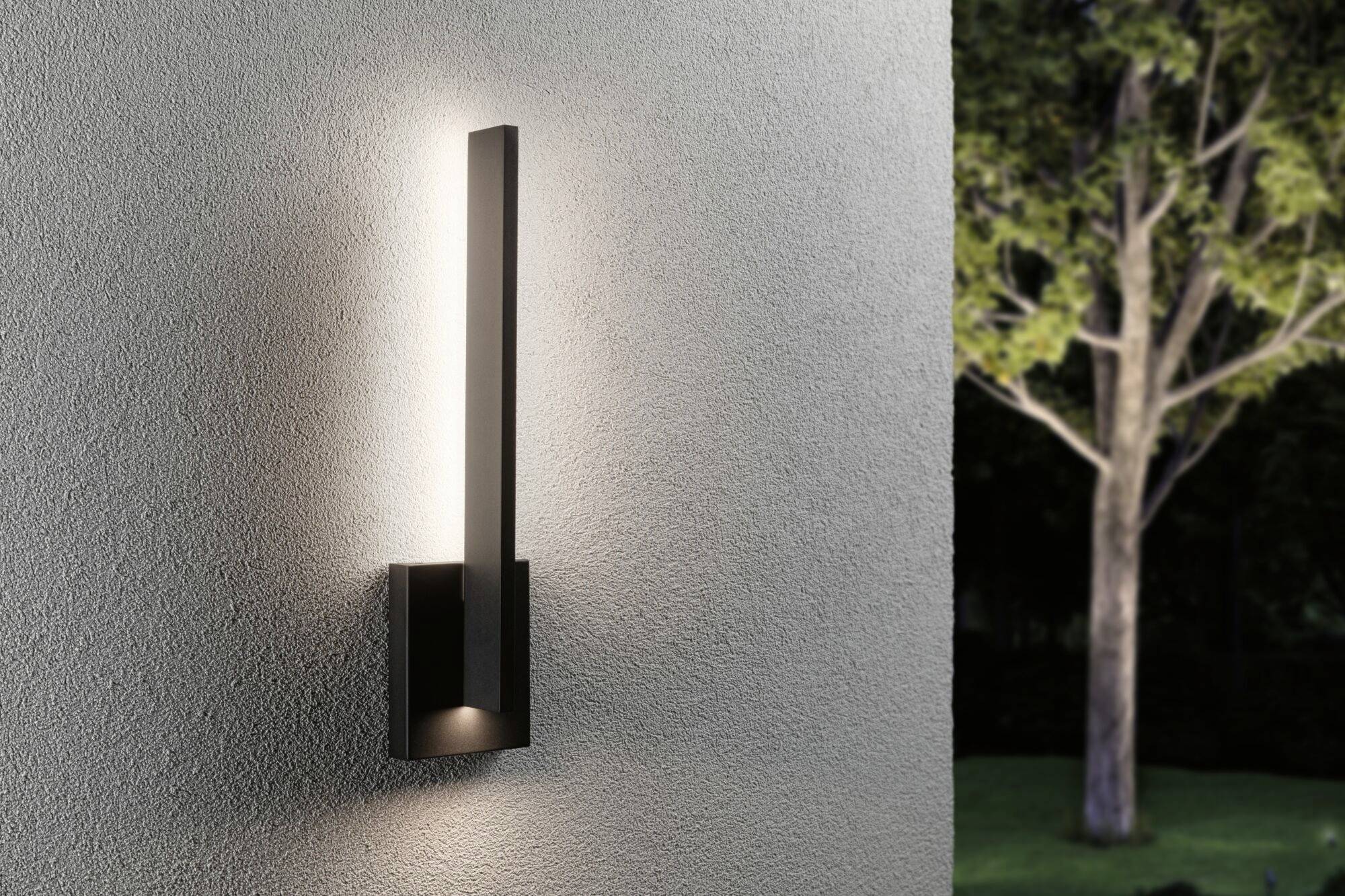 Een moderne buitenwandlamp met verticaal, rechthoekig ontwerp verlicht een grijze muur. Op de achtergrond zijn bomen zichtbaar.