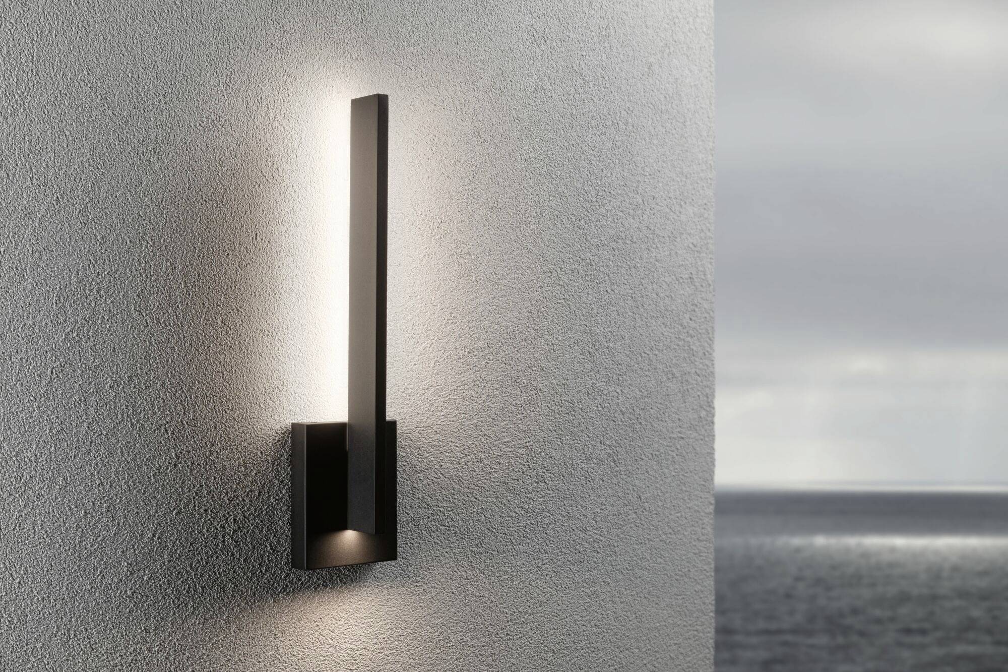 Moderne zwarte wandlamp straalt warm licht tegen een grijze muur. Op de achtergrond zee en een bewolkte lucht.