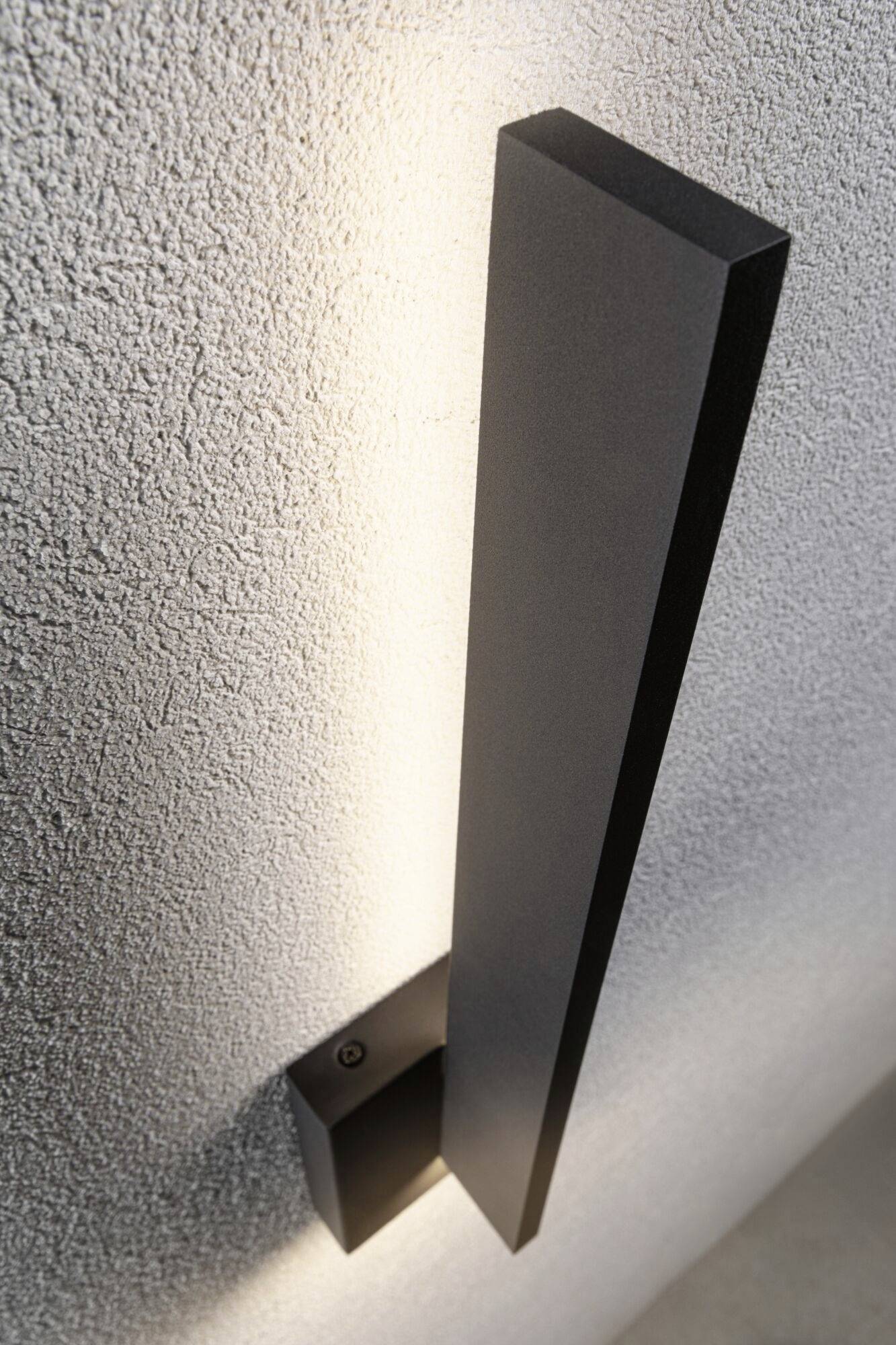 Een moderne, zwarte wandlamp aan een gestructureerde grijze oppervlakte, die zacht licht naar boven uitstraalt.