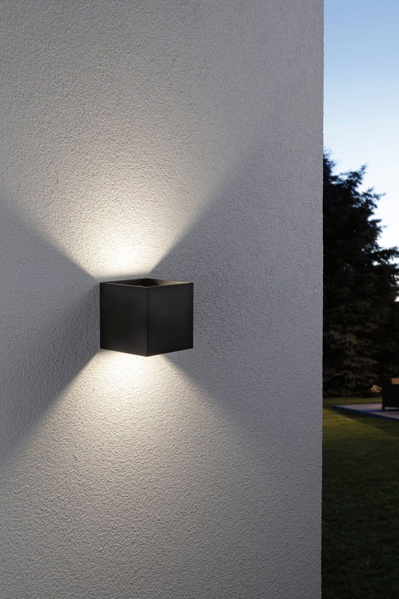 Een kubische wandlamp straalt zacht licht in X-vorm tegen een witte muur. Op de achtergrond is een boom zichtbaar bij het invallen van de avond.