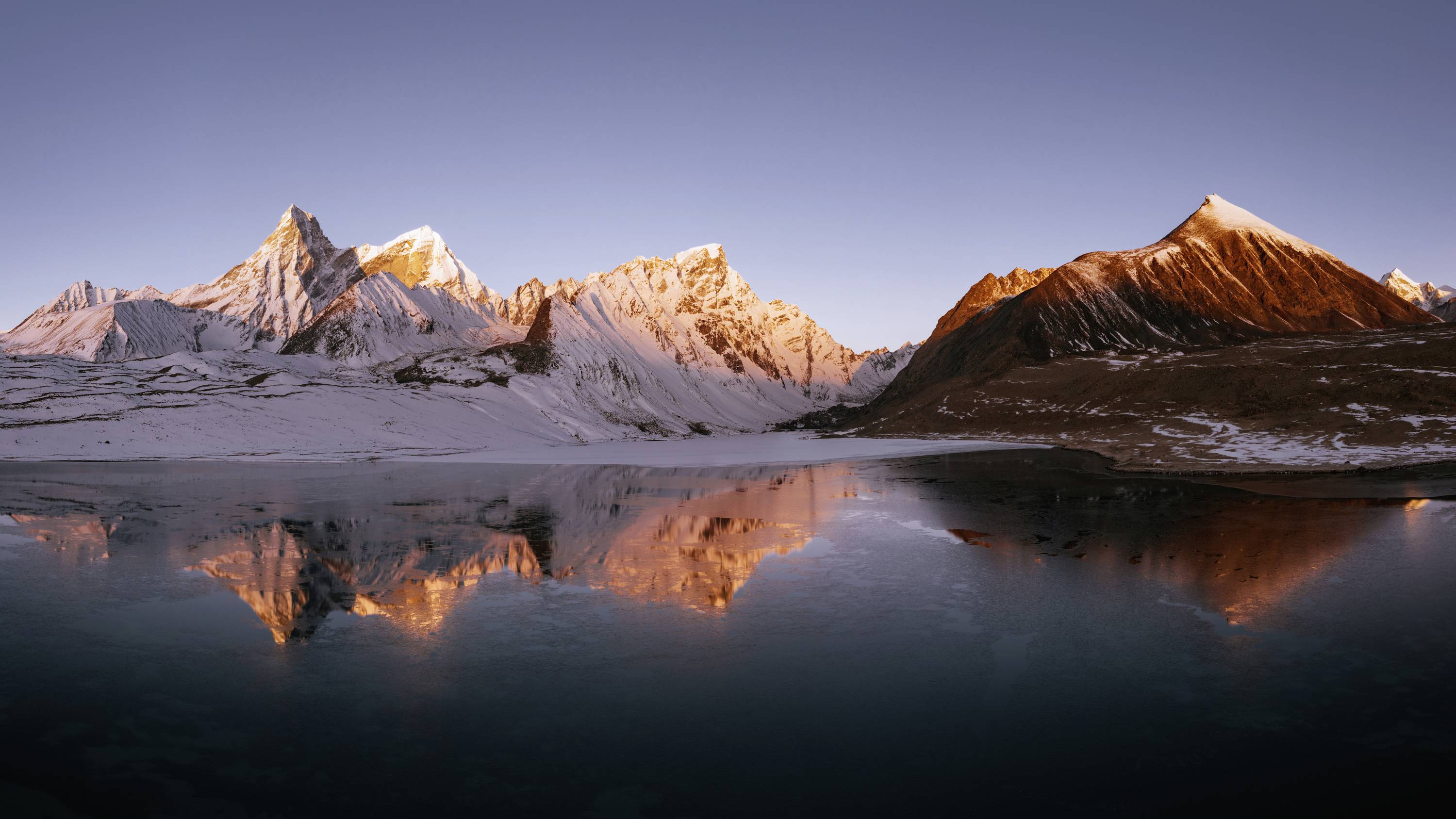 Bergpanorama met met sneeuwbedekte toppen en een rustige meer op de voorgrond, weerspiegelt het warme licht van de ondergaande zon.