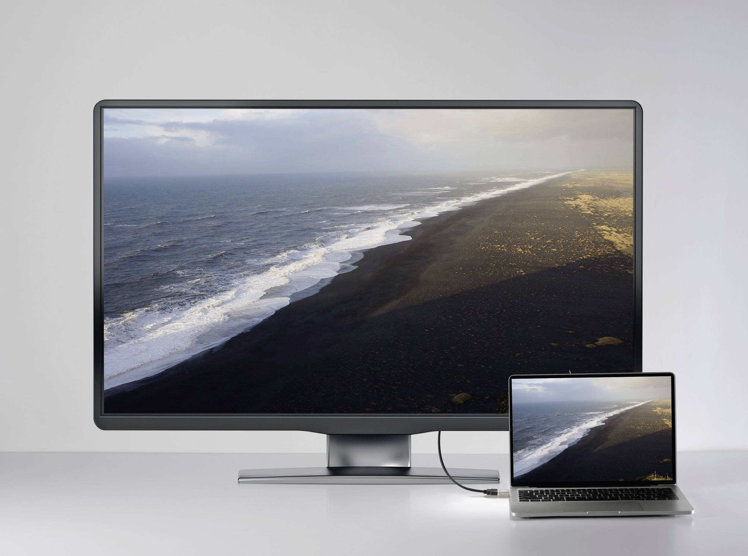 Een grote monitor toont een afbeelding van een kustrand met donker zand en golven. Naast de monitor is een laptop aangesloten die hetzelfde beeld vertoont.