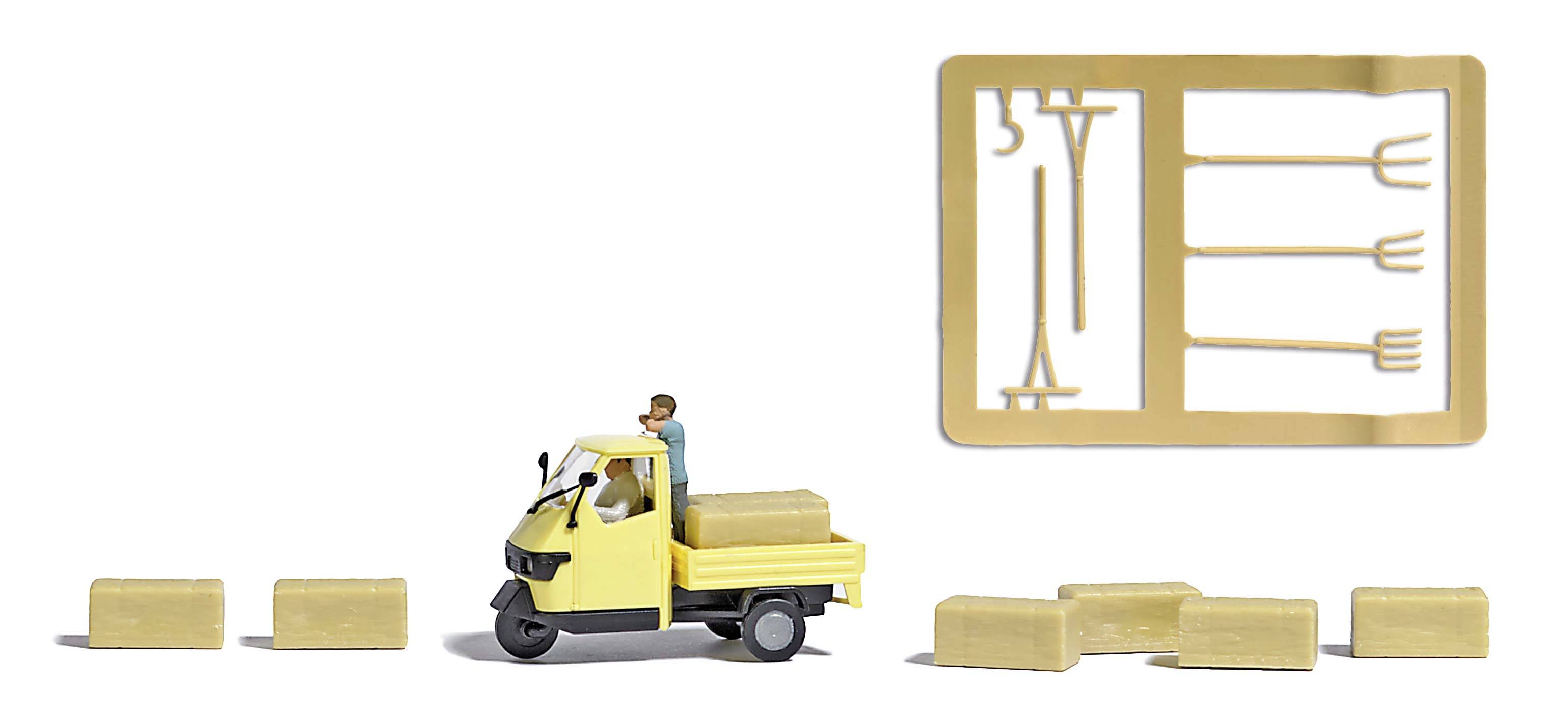 Een miniatuurfiguur rijdt een geel driewielig voertuig en vervoert hooibalen. Rechts daarnaast een gietframe met gereedschappen.