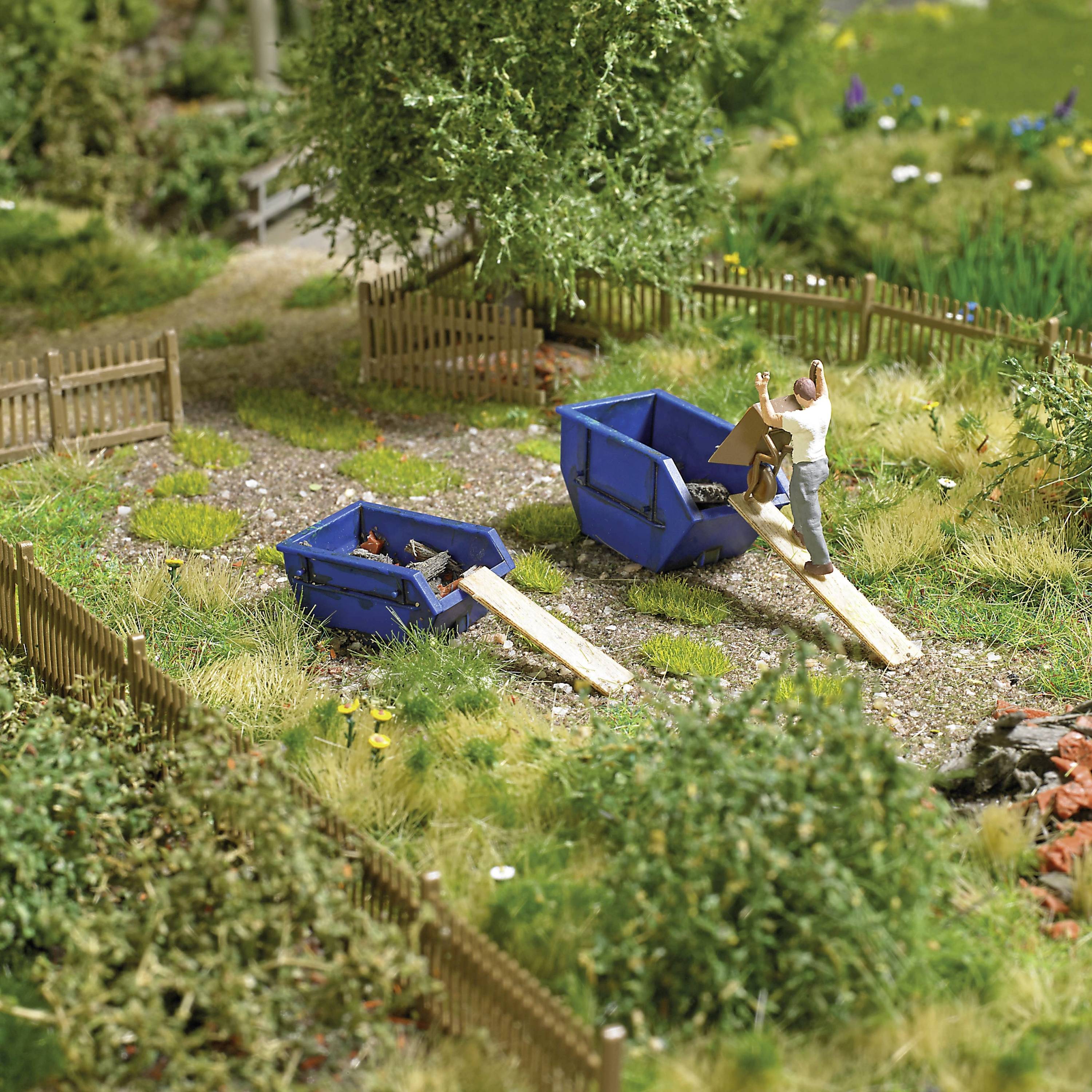 Een miniatuurtafereel toont twee personen die puin storten in blauwe containers naast een kleine, omheinde tuinzone.