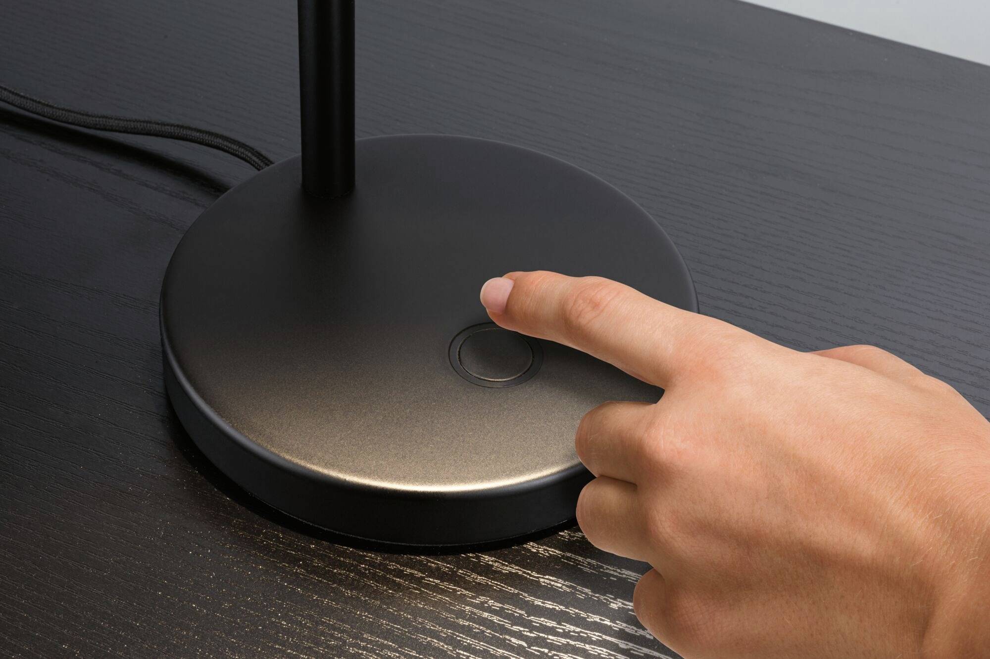 Een hand raakt de aanraakschakelaar aan de voet van een tafellamp. De lamp staat op een donkere houten tafel.