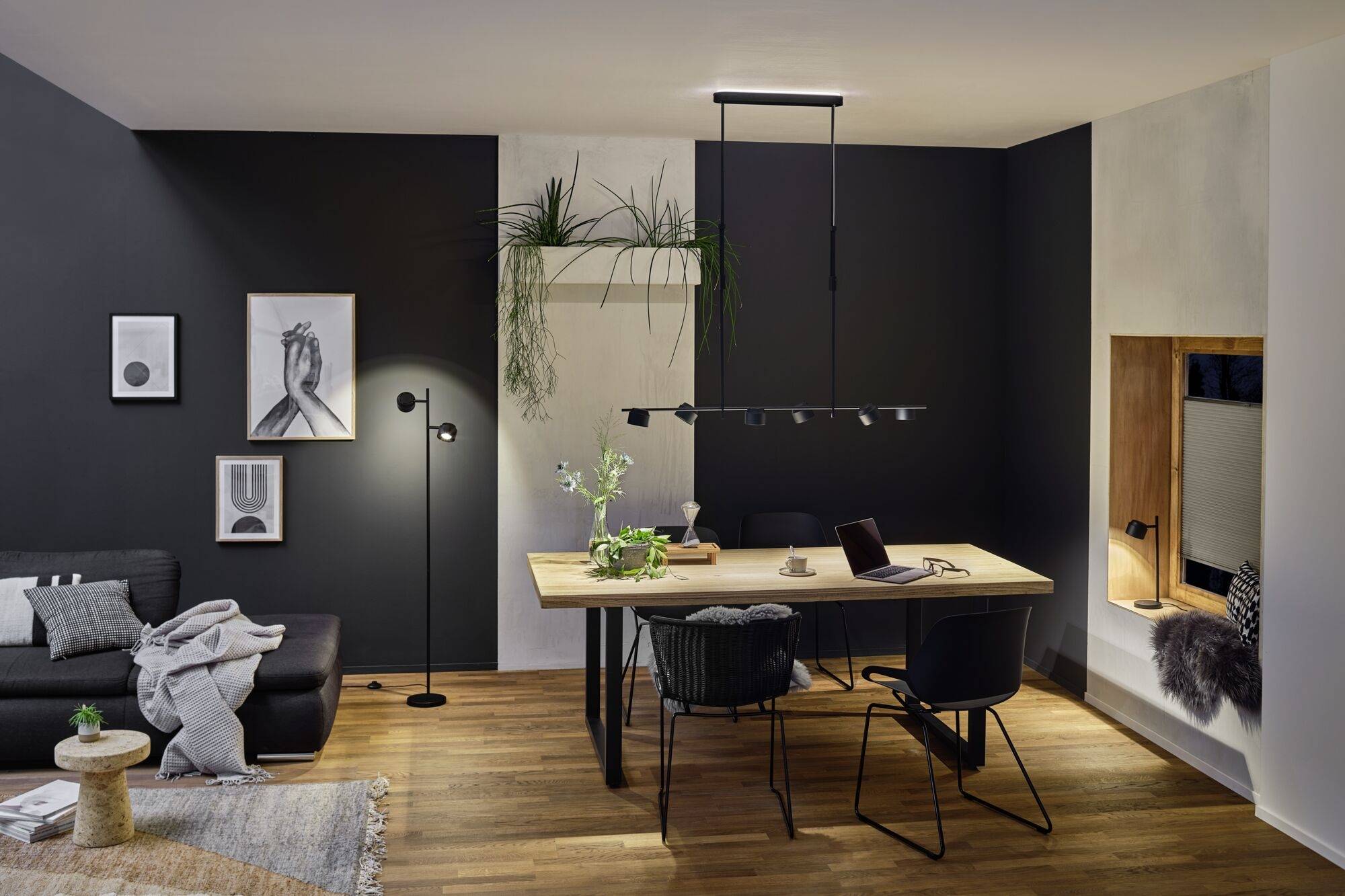 Modern woonkamer met eettafel en stoelen, een stijlvolle zwarte bank, decoratieve schilderijen aan de muur en hangende planten.