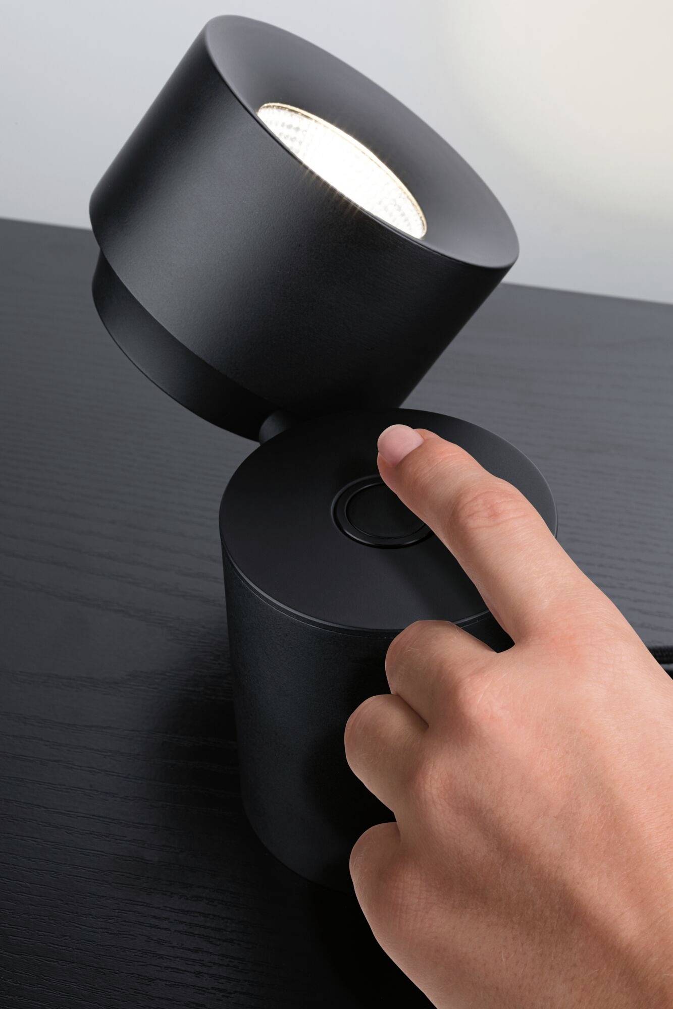 Een hand drukt op een schakelaar van een moderne, zwarte lamp op een tafel. De lamp is kantelbaar en verlicht.