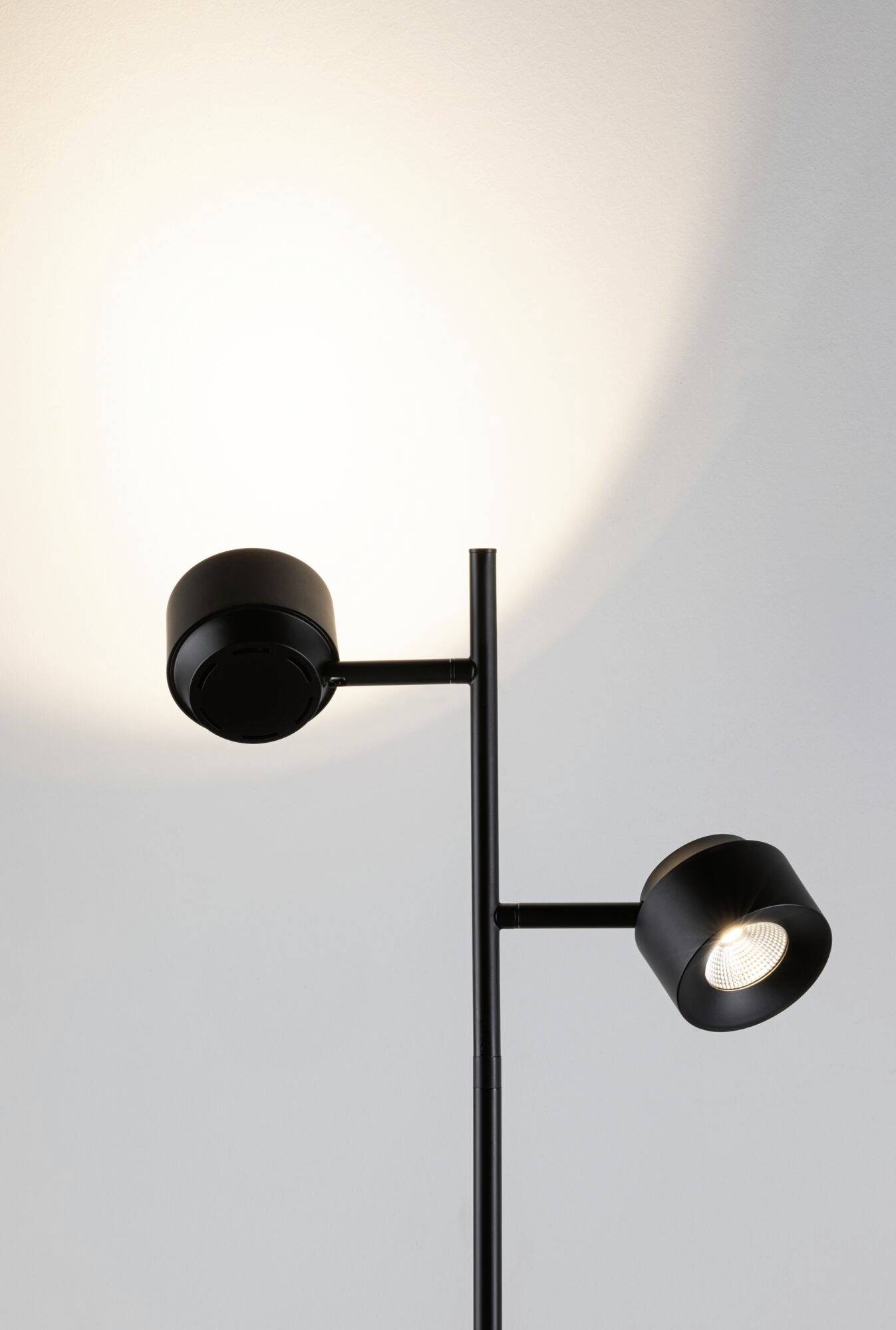 Zwarte vloerlamp met twee verstelbare spots die zacht, warm licht tegen de muur werpen. Minimalistisch design.