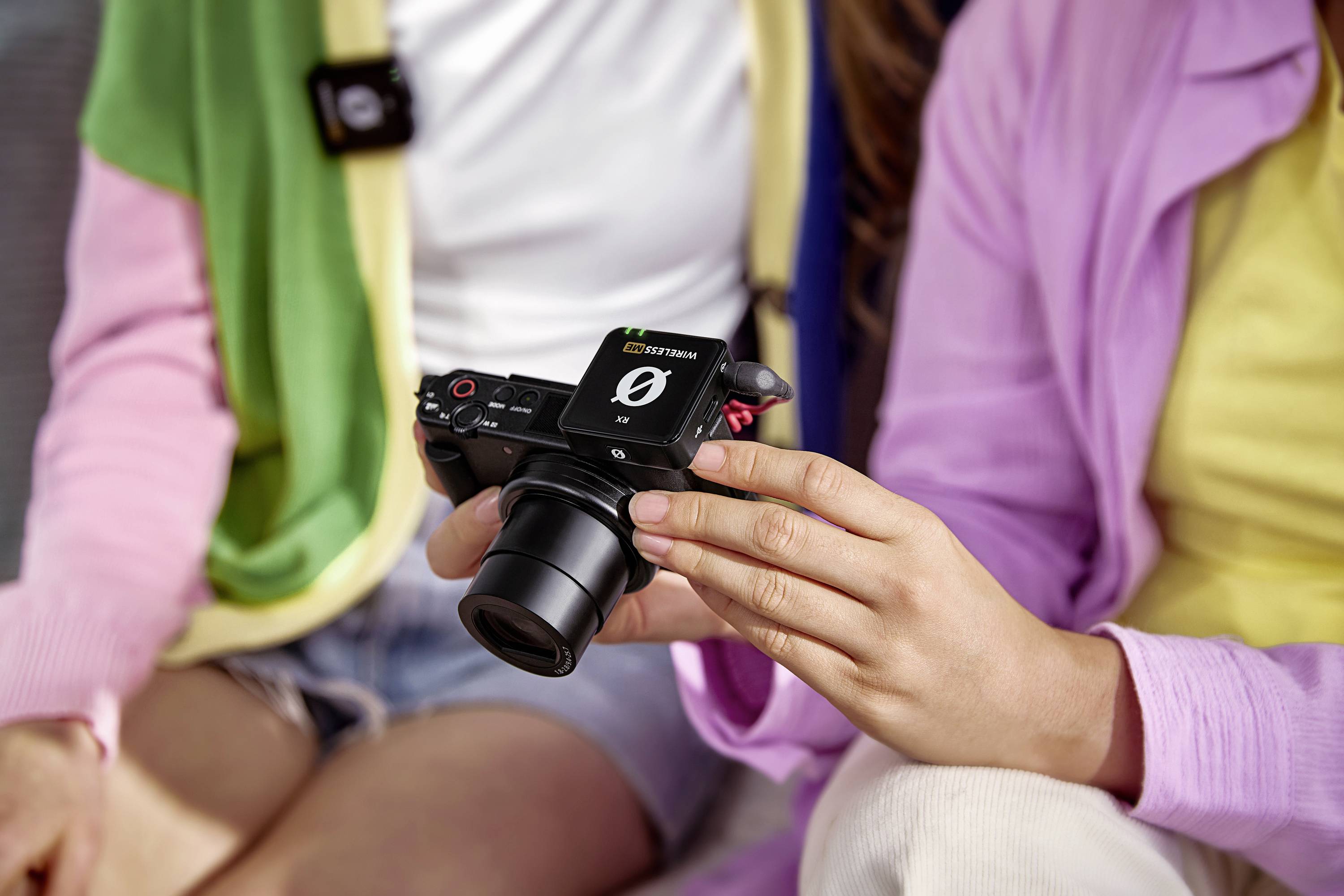 Een persoon houdt een camera vast terwijl een andere persoon naast hem zit. Beide dragen kleurrijke kleding. De scène oogt ontspannen en is gericht op de camera.