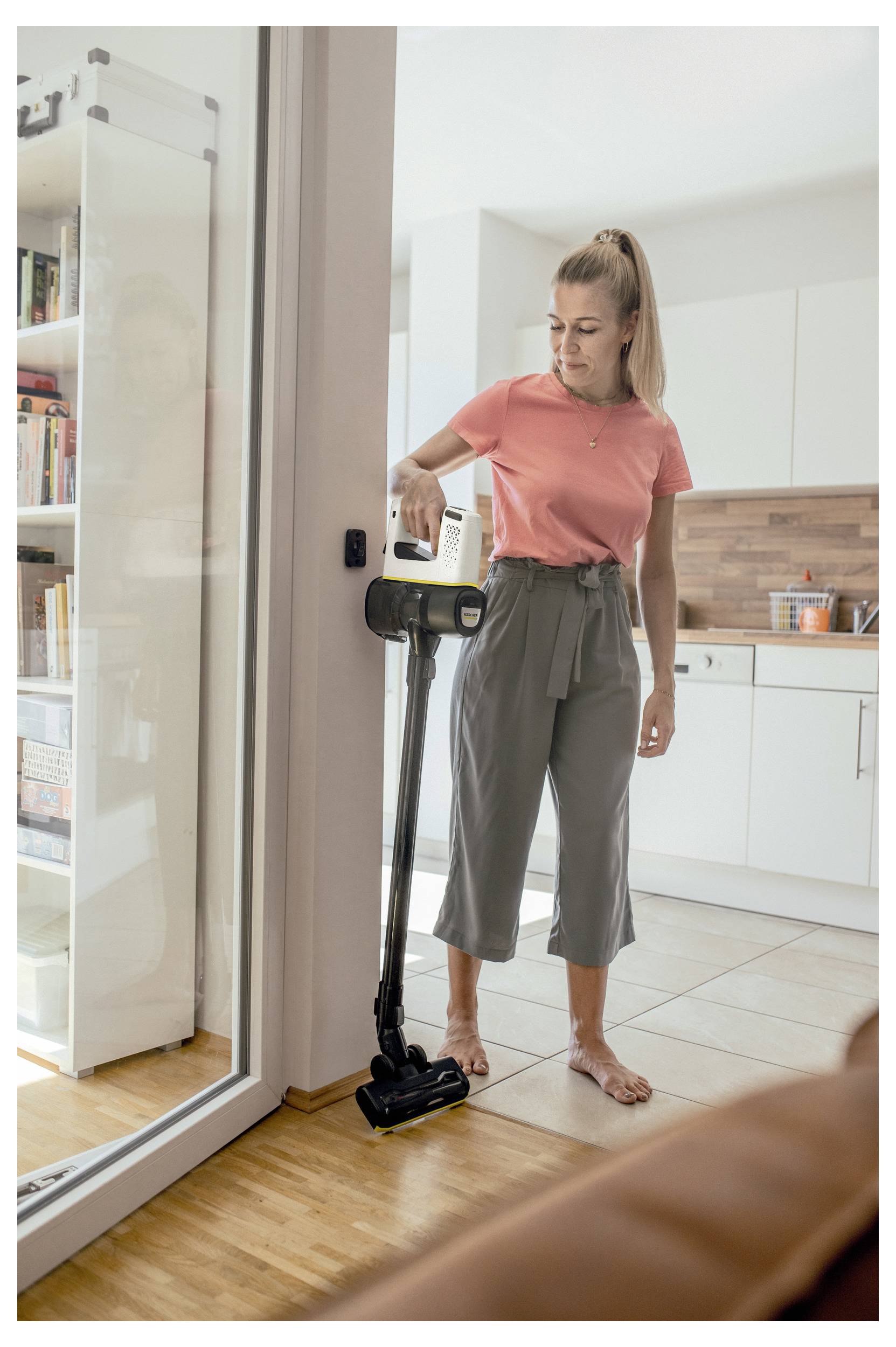 Persoon in een roze shirt die de woonkamer stofzuigt met een draadloze steelstofzuiger. Lichte, heldere binnenruimte, met de keuken op de achtergrond.