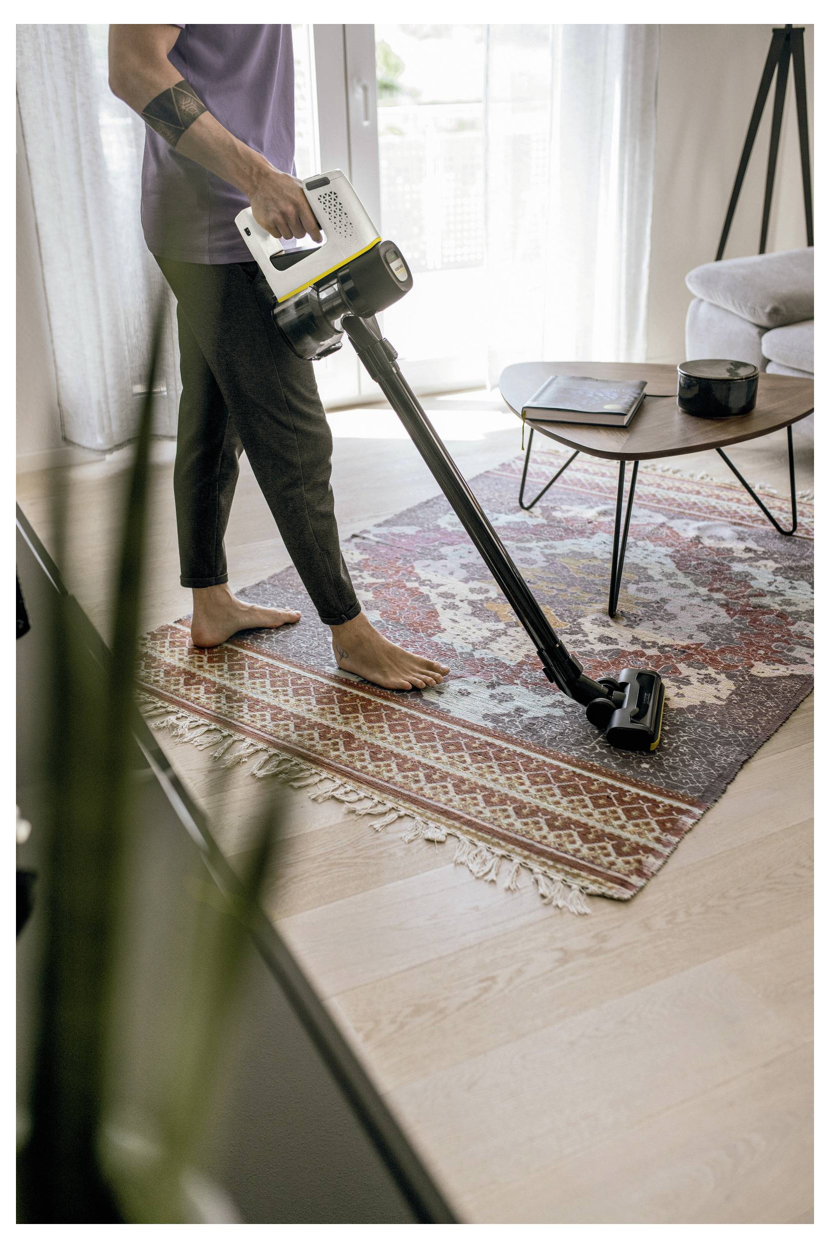 Een persoon stofzuigt een gedessineerd vloerkleed in een woonkamer met een snoerloze stofzuiger. In de buurt staat een kleine tafel met enkele voorwerpen bovenop.