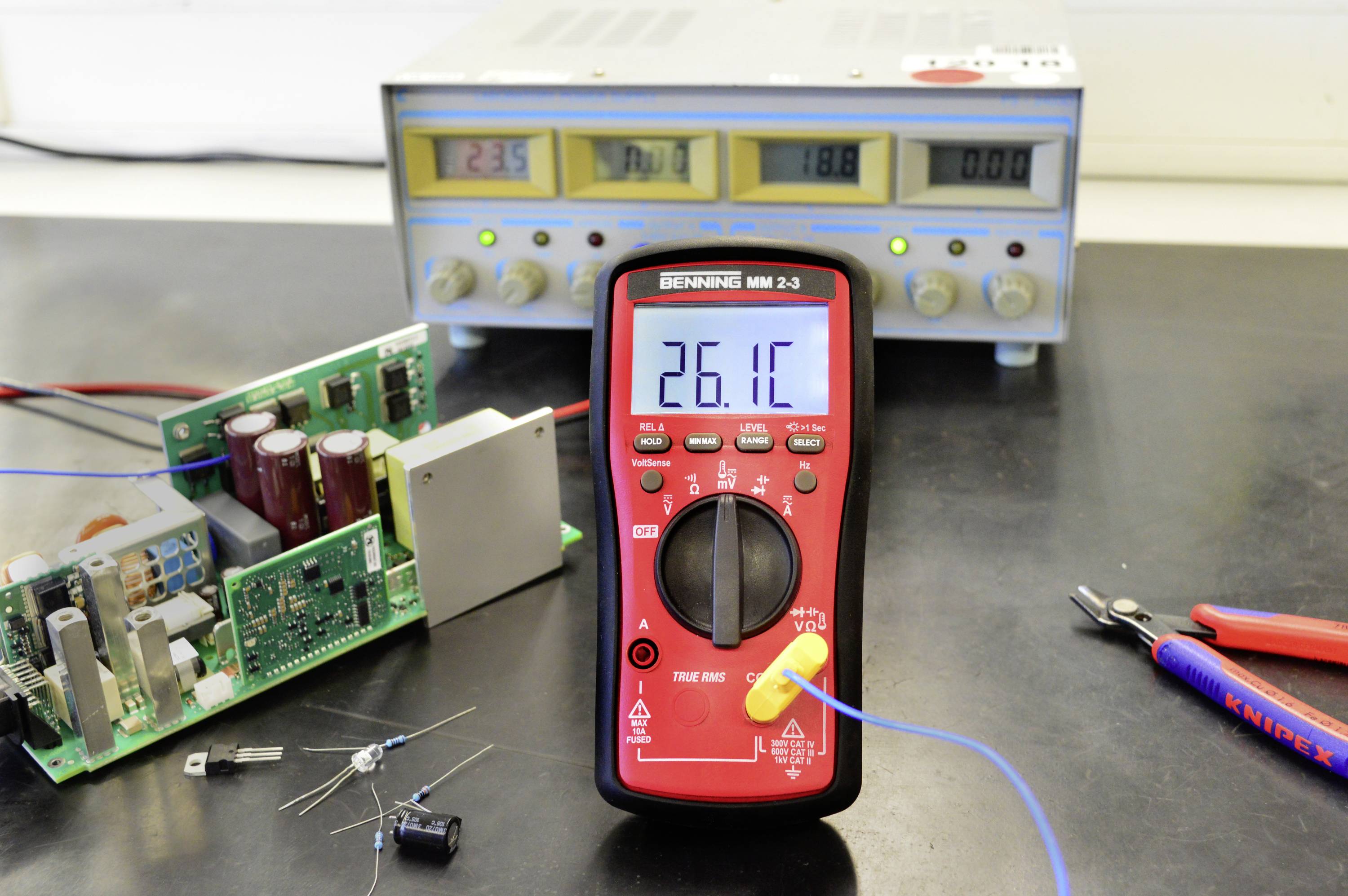 Een digitale multimeter geeft 26,1 graden aan. Op de achtergrond staat een laboratoriumvoedingsapparaat met verschillende stroomsterktes en spanningen.