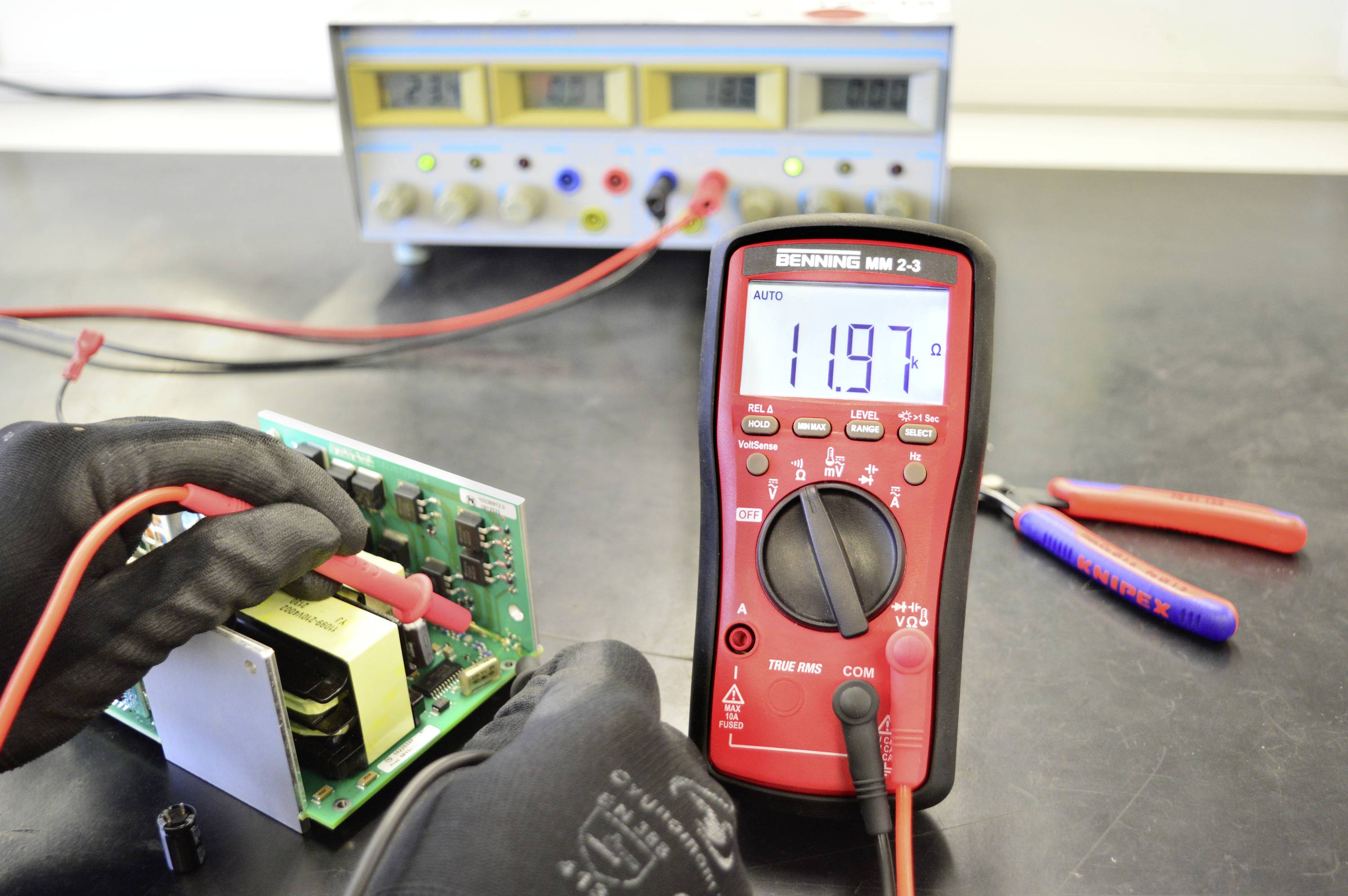 Een technicus meet de spanning op een printplaat met een multimeter die 11,97 volt aangeeft. Op de achtergrond staat een voedingsapparaat.
