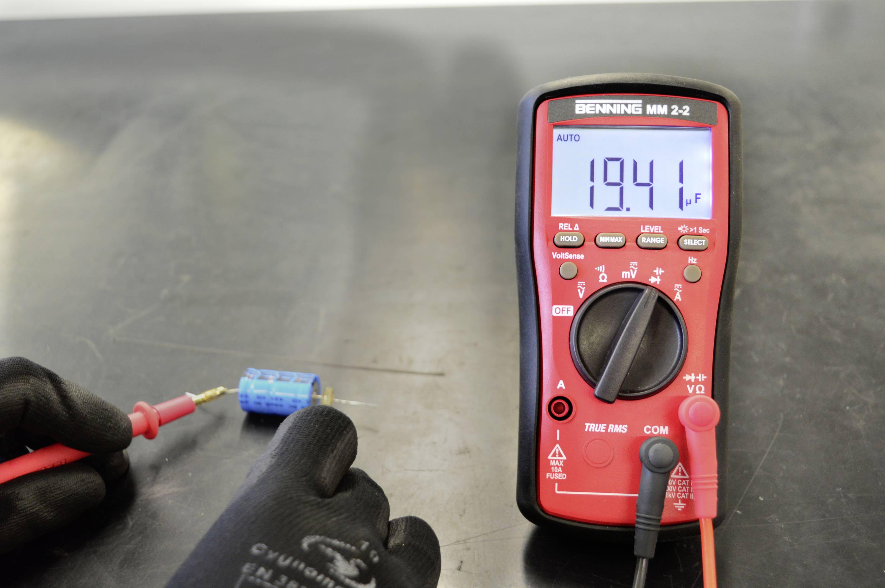 Een rode multimeter toont '19,41' op een digitaal display. Een hand houdt een testprobe vast die is aangesloten op een blauw elektronisch onderdeel.