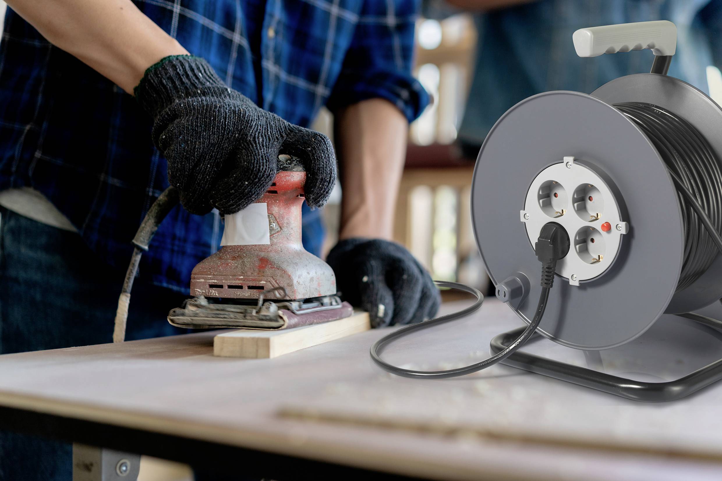 Een persoon schuurt hout met een schuurmachine op een tafel. Op de voorgrond staat een kabelhaspel met meerdere stopcontacten.