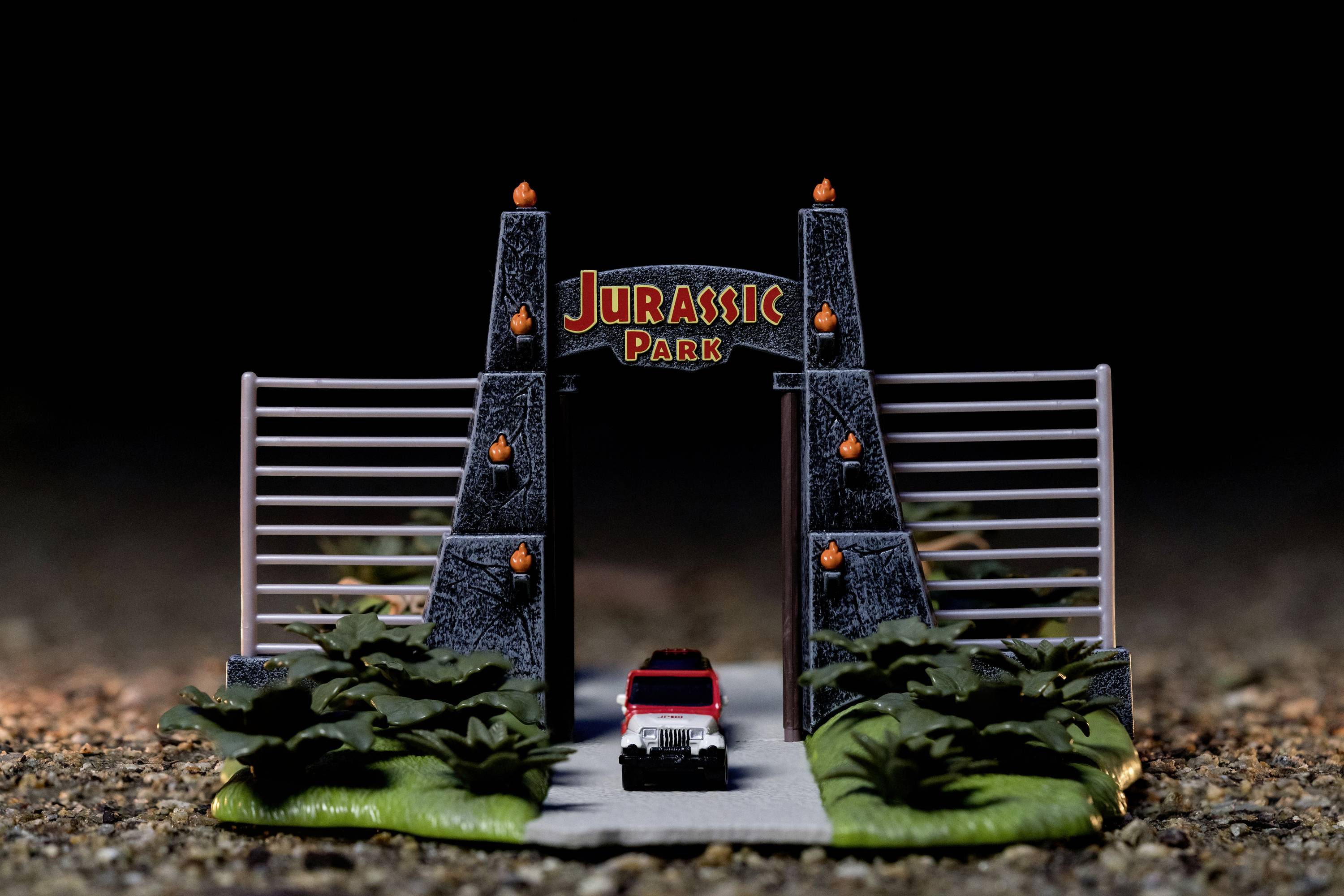 Een kleine auto rijdt door een miniatuurpoort met de tekst 'Jurassic Park', omgeven door planten, op een rotsachtige ondergrond.