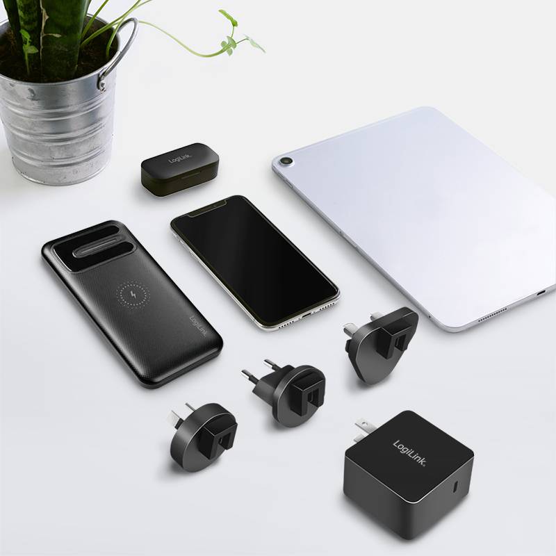 Verschillende elektronische apparaten, waaronder een smartphone, tablet, laadadapter en stekkerdoos-adapter op een tafel naast een plant.