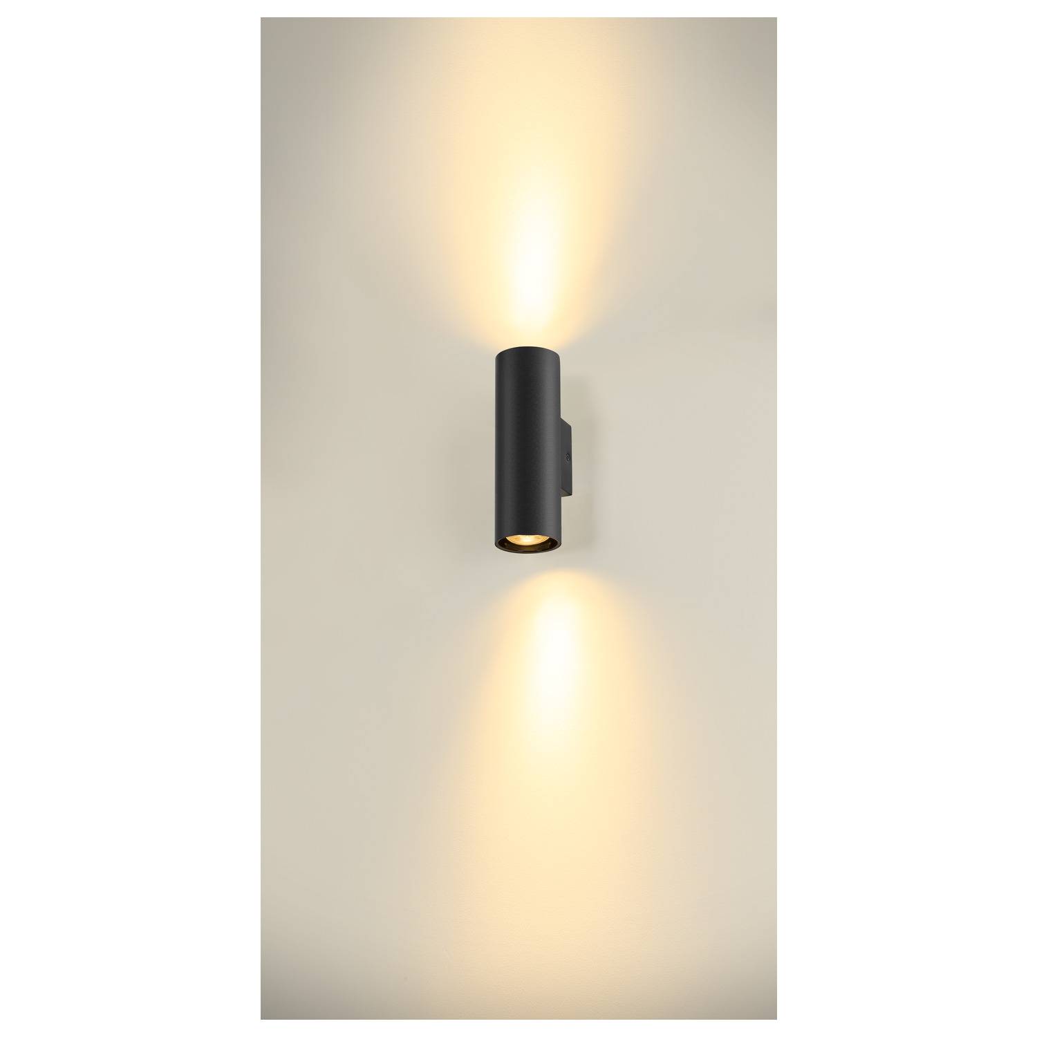Zwarte wandlamp met cilindrisch design. Hij straalt warm licht zowel naar boven als naar beneden tegen een lichte muur.