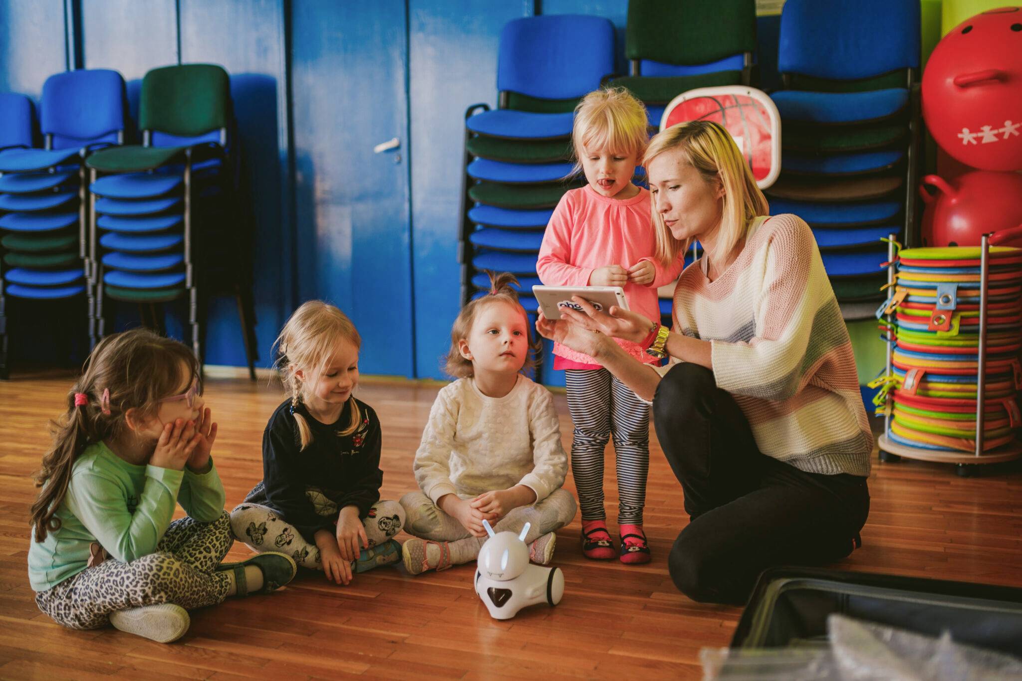 Een volwassene toont een tablet aan vier kinderen in een ruimte. Een kleine speelgoedrobot staat op de vloer. De kinderen zien er geïnteresseerd uit.