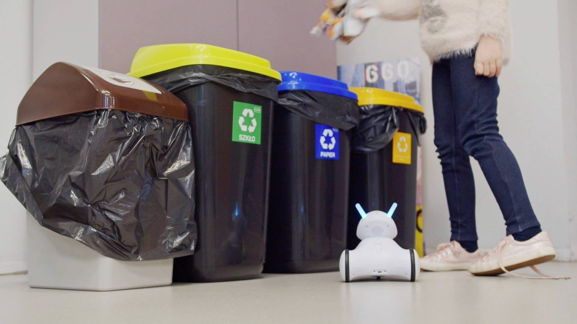 Een persoon gooit afval in gekleurde recyclingcontainers voor glas, plastic en papier. Een kleine robot zit op de grond.