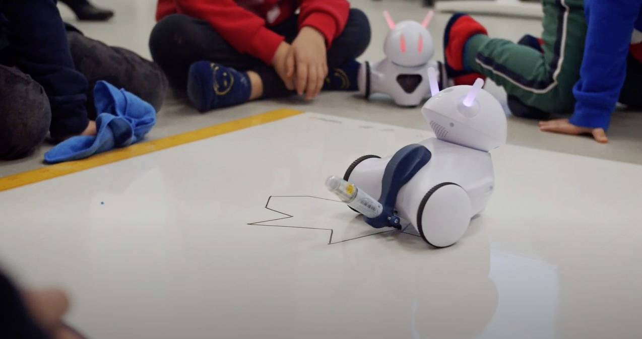 Kinderen observeren een kleine robot op wielen die zich over de vloer beweegt en een lijn trekt. Een andere robot staat op de achtergrond.