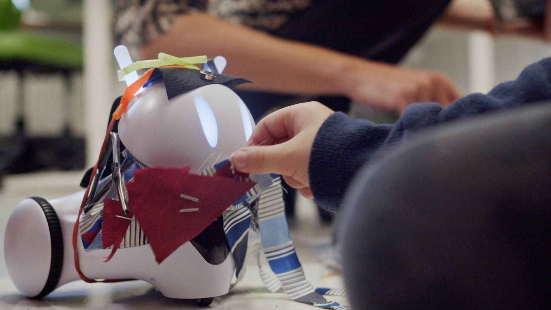 Een kind versiert een kleine robot met kleurrijke stofstroken. Op de achtergrond is een persoon te zien die de robot bekijkt.