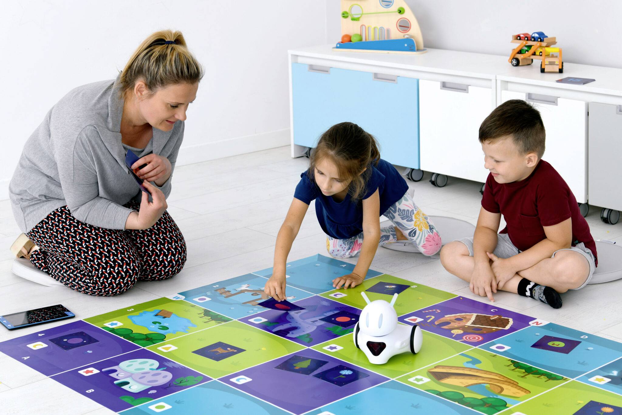 Een vrouw en twee kinderen spelen met een vloerrobot op een kleurrijke mat die verschillende activiteiten en afbeeldingen toont.