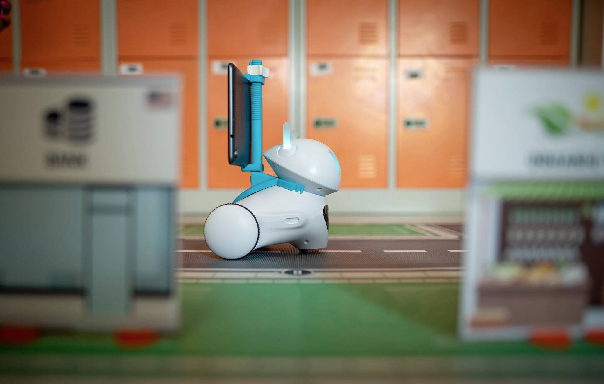 Een kleine, blauwe robot rijdt op een weg tussen miniatuurbouwen. Op de achtergrond zijn oranje kluisjes zichtbaar.