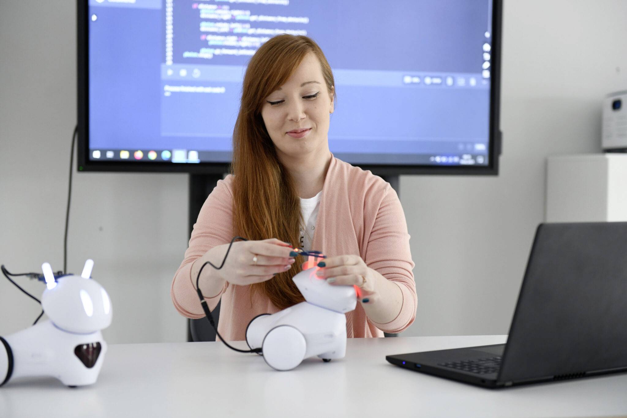 Een vrouw programmeert robots naast een laptop. Op de achtergrond is een scherm met code zichtbaar.