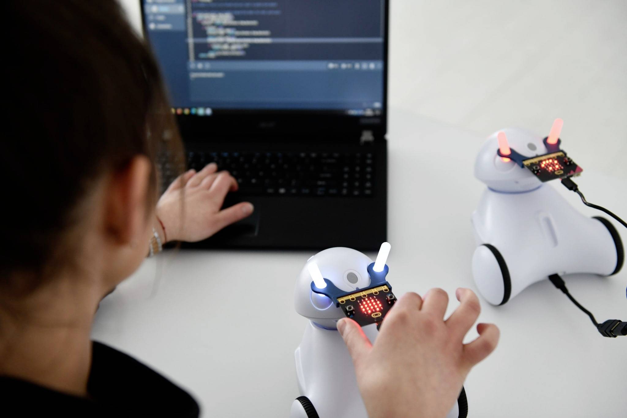 Een persoon programmeert robots met een laptop. Twee witte robots met verlichte displays staan op een tafel.