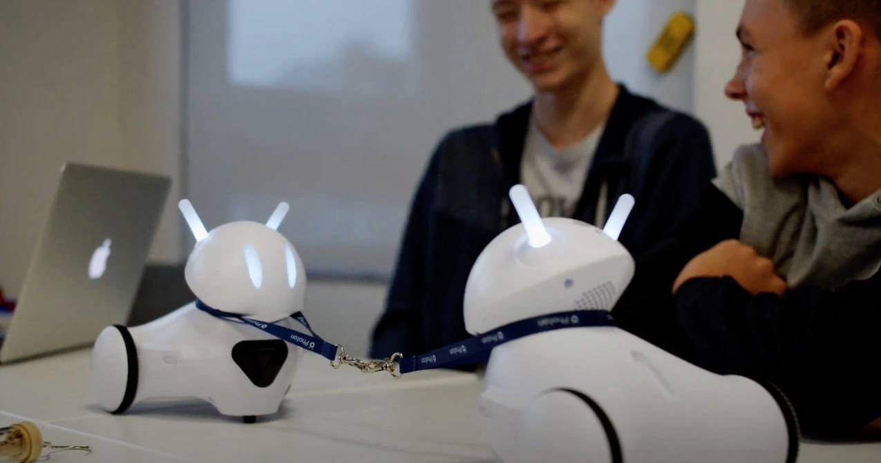 Twee personen zitten aan een tafel en kijken glimlachend naar twee kleine, witte robots met oplichtende oren die met een band met elkaar verbonden zijn.