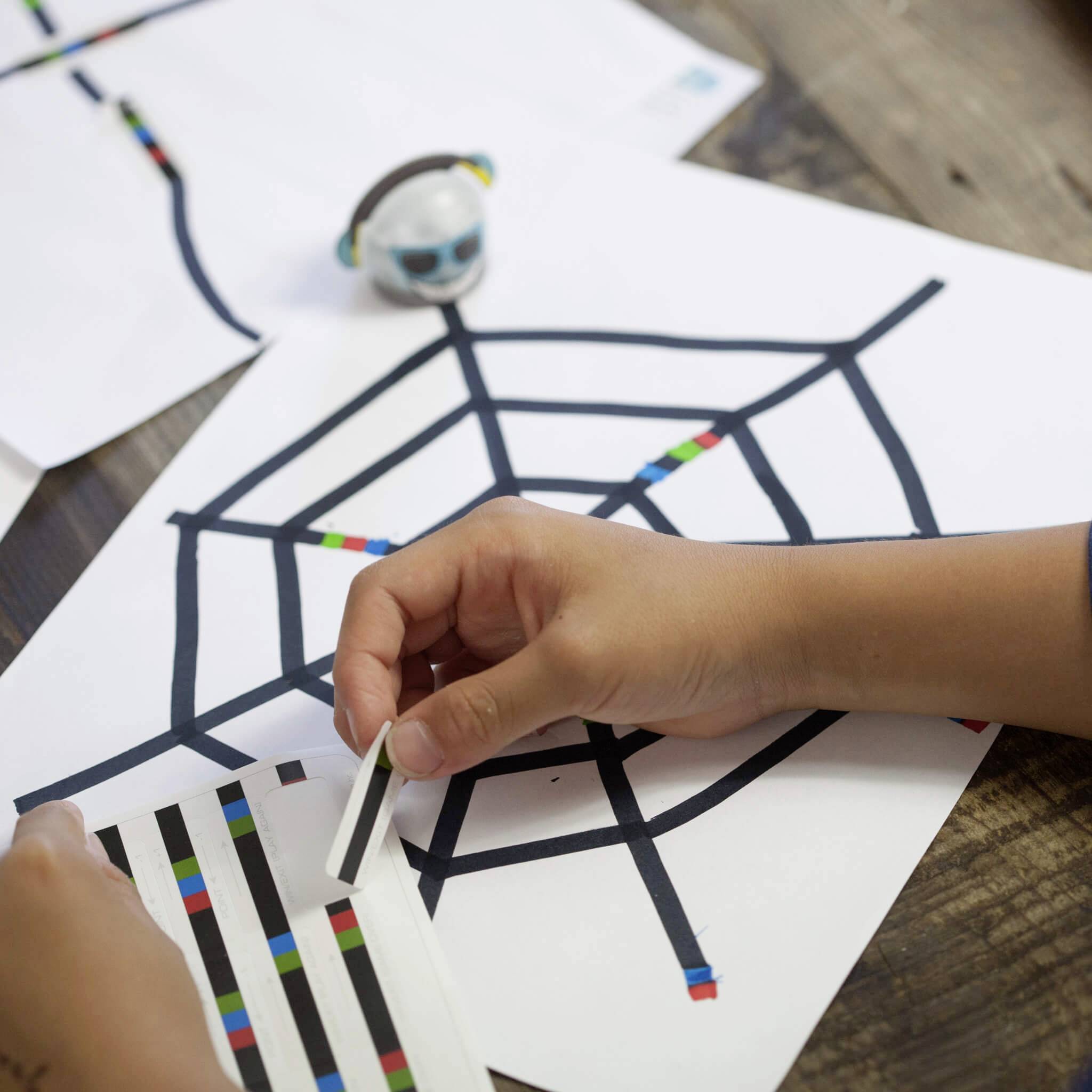 Een persoon plakt gekleurde strepen op een papier met lijnen, blijkbaar onderdeel van een knutselproject op een houten tafel.