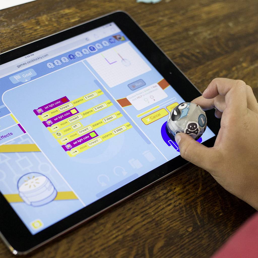 Een tablet toont een programmeer-app voor kinderen. Een hand bestuurt een kleine robot over het scherm.