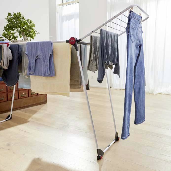 Een droogrek met kleurrijke kleding staat naast een raam. Op de achtergrond is een plant op een kast te zien.