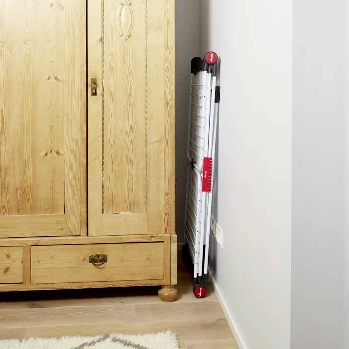 Een houten kast staat op een houten vloer. Ernaast leunt een ingeklapte droogrek tegen de muur.