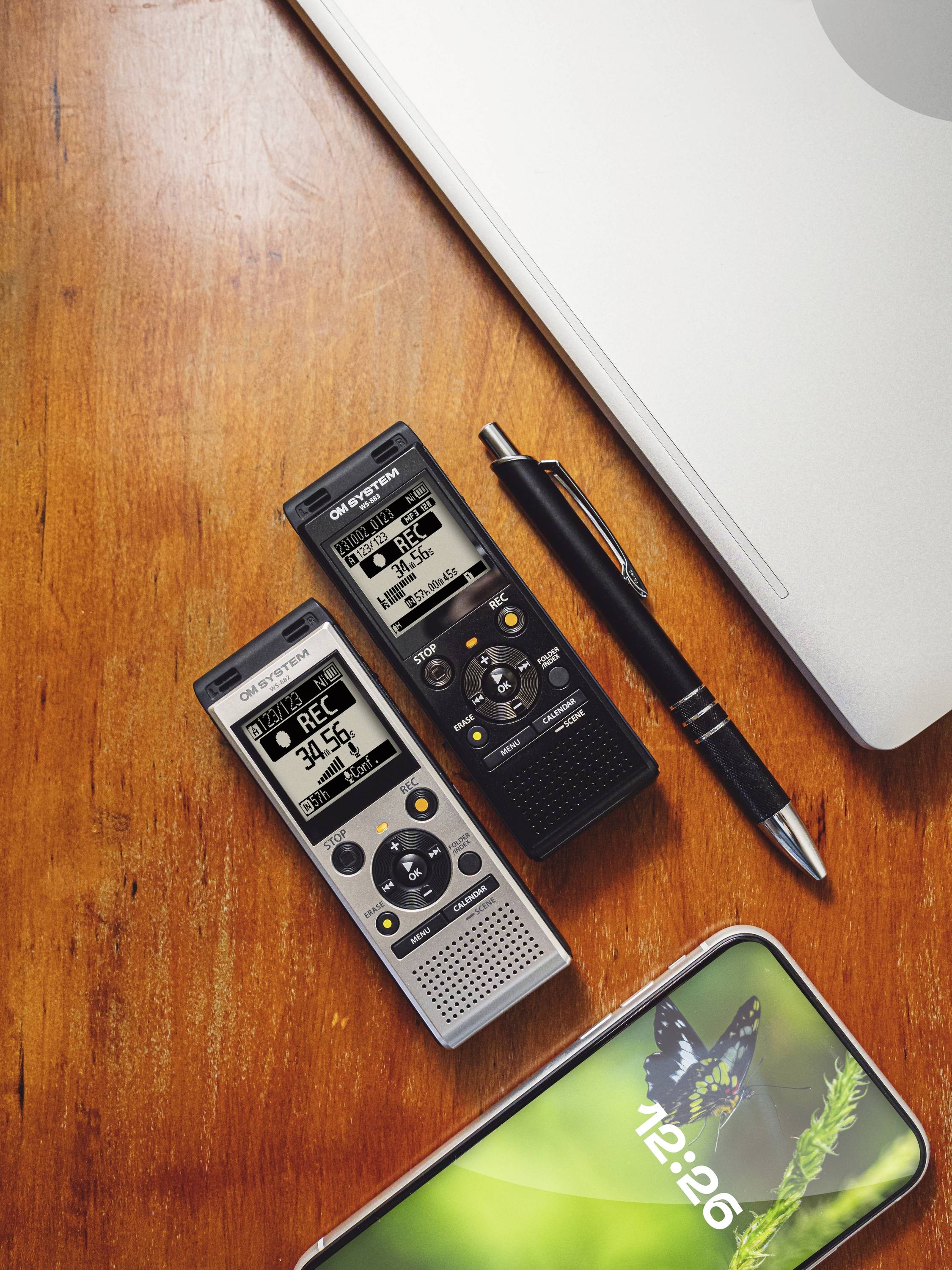 Twee digitale dicteerapparaten, een pen en een smartphone op een houten tafel naast een laptop. De smartphone toont een vlinderplaatje.