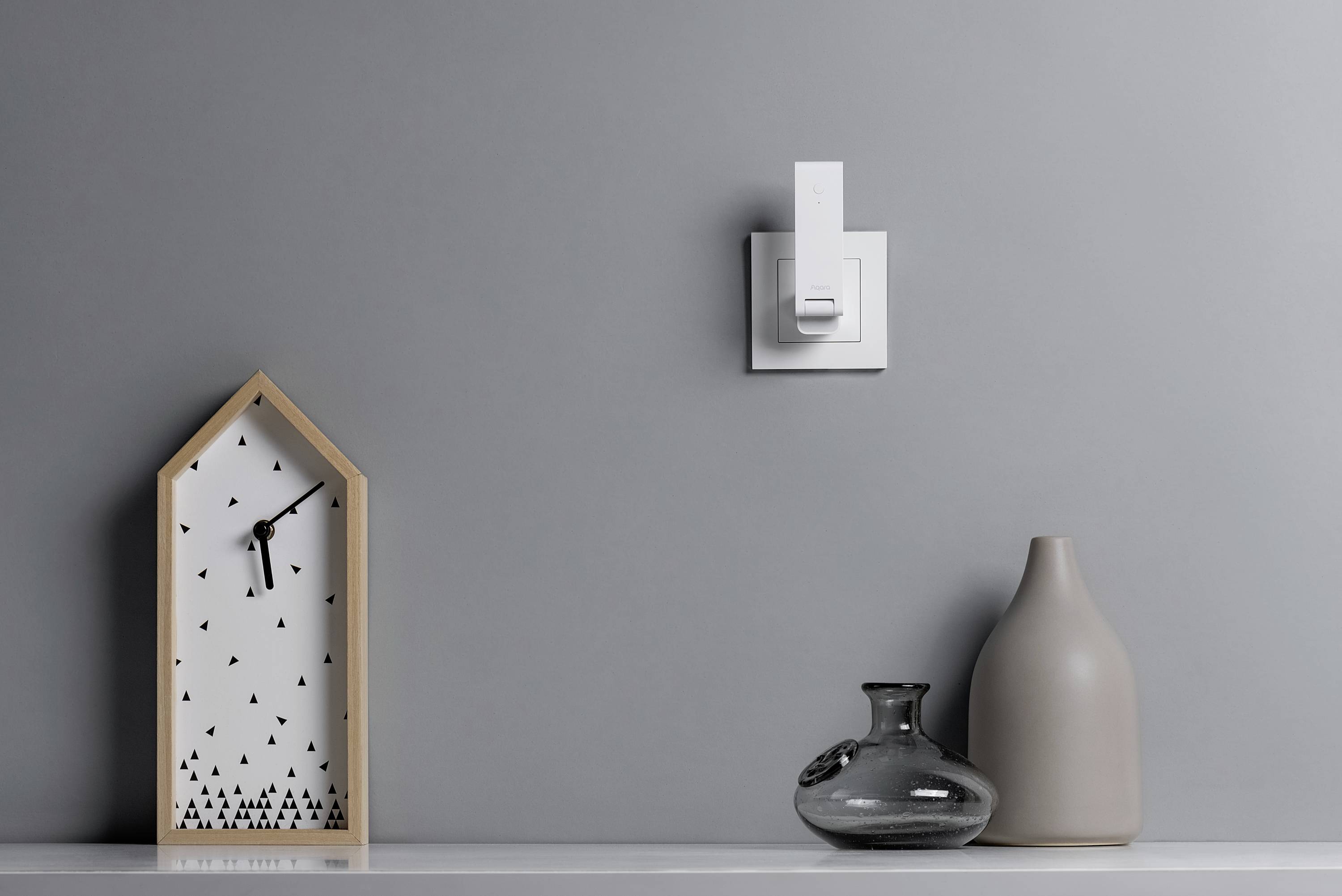 Houten klok in huisvorm met zwarte wijzerplaat links, drie vazen rechts op een plank voor een grijze muur met een witte lichtschakelaar.