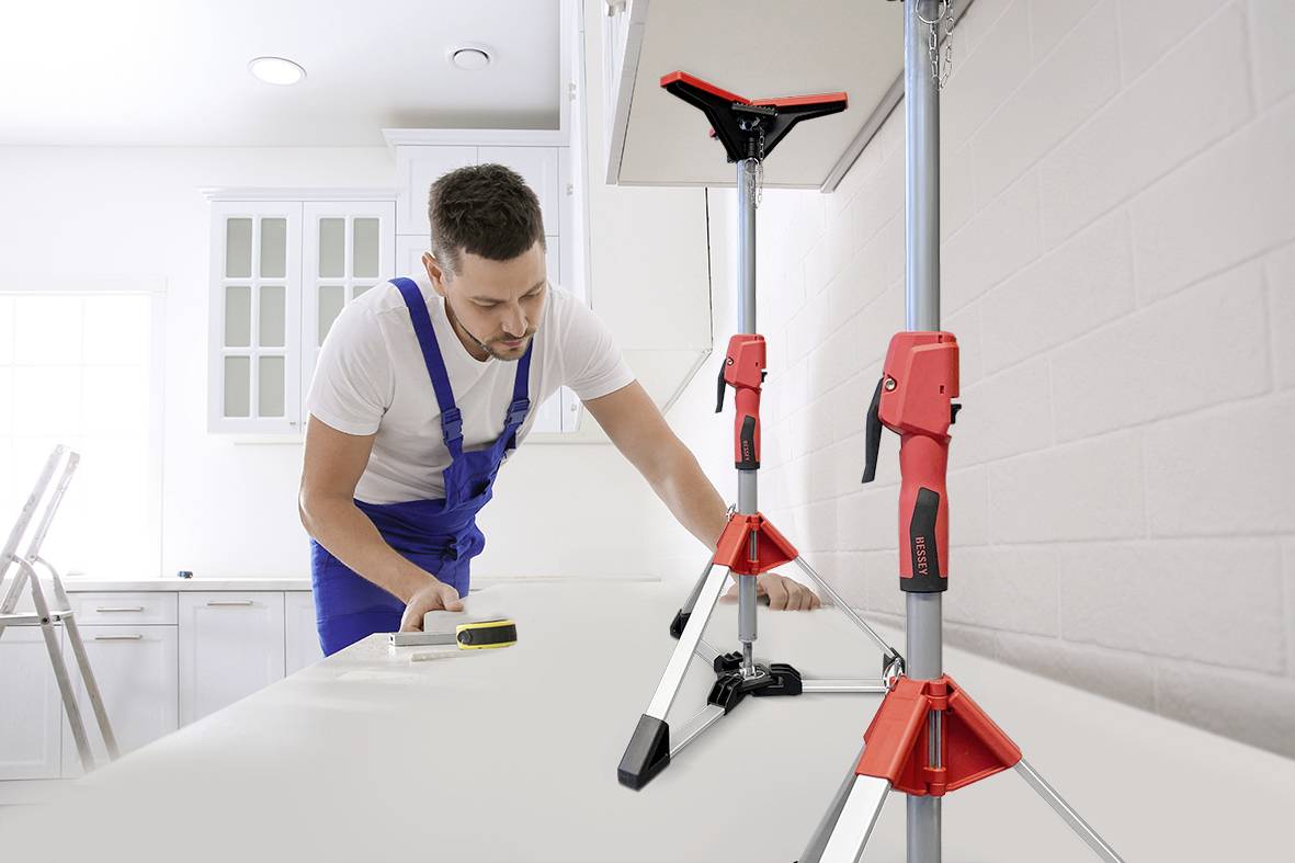 Een vakman in blauwe werkkleding meet een werkblad in een moderne keuken. Twee rode steunen ondersteunen de keukenkastjes.