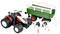 Een speelgoed tractor met aanhanger naast een koeienfiguur. De tractor is rood-wit, de aanhanger groen, en de koe zwart-wit.