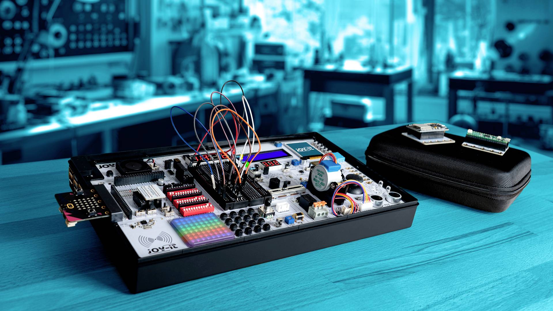 Elektronische set met kabels en componenten op een houten tafel, met een onscherpe werkplaatsachtergrond; toont leerapparatuur voor elektrotechniek.