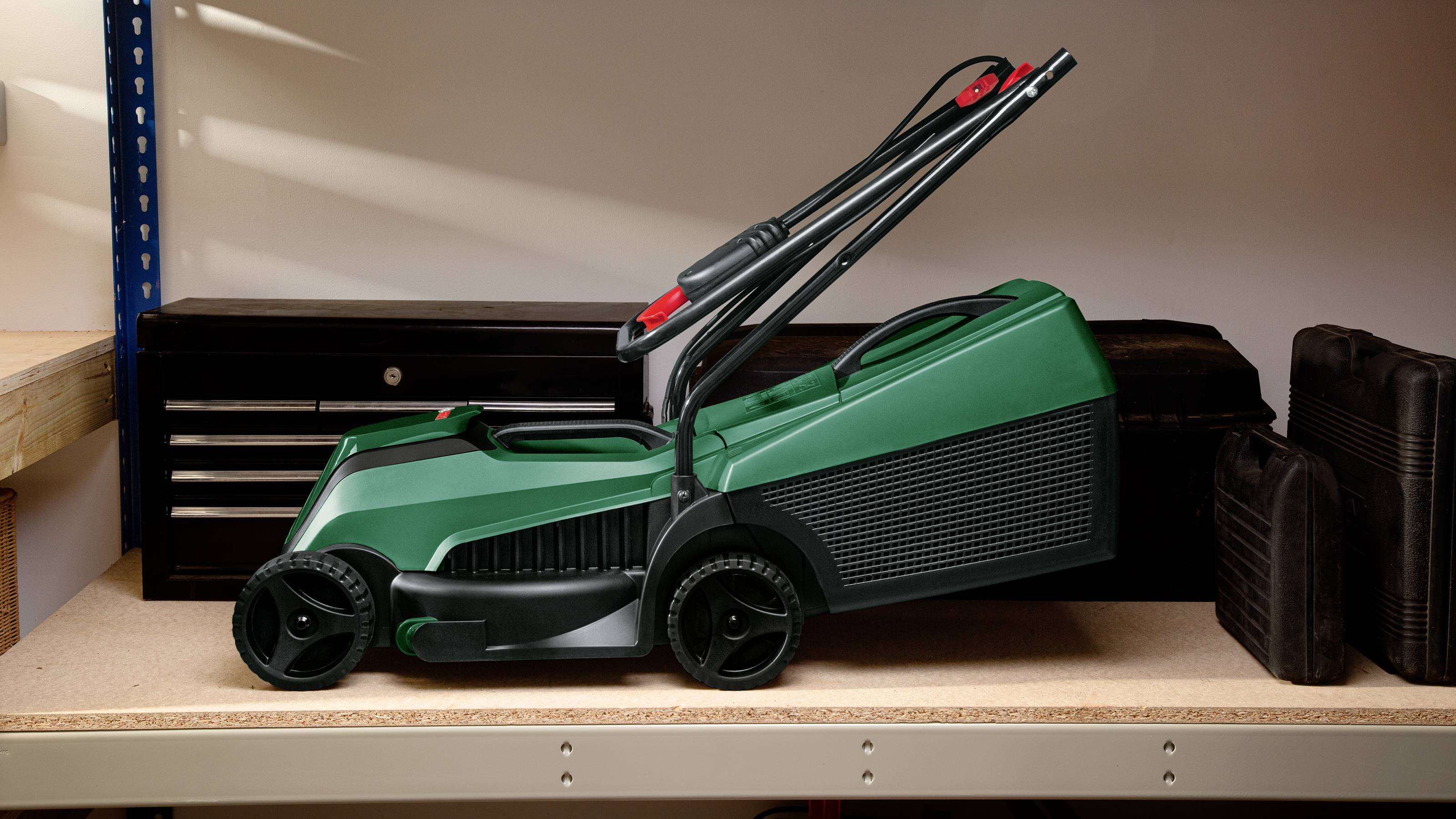 Een groene grasmaaier staat op een tafel in een werkplaats, omringd door gereedschapskisten. Het handvat is naar beneden geklapt.