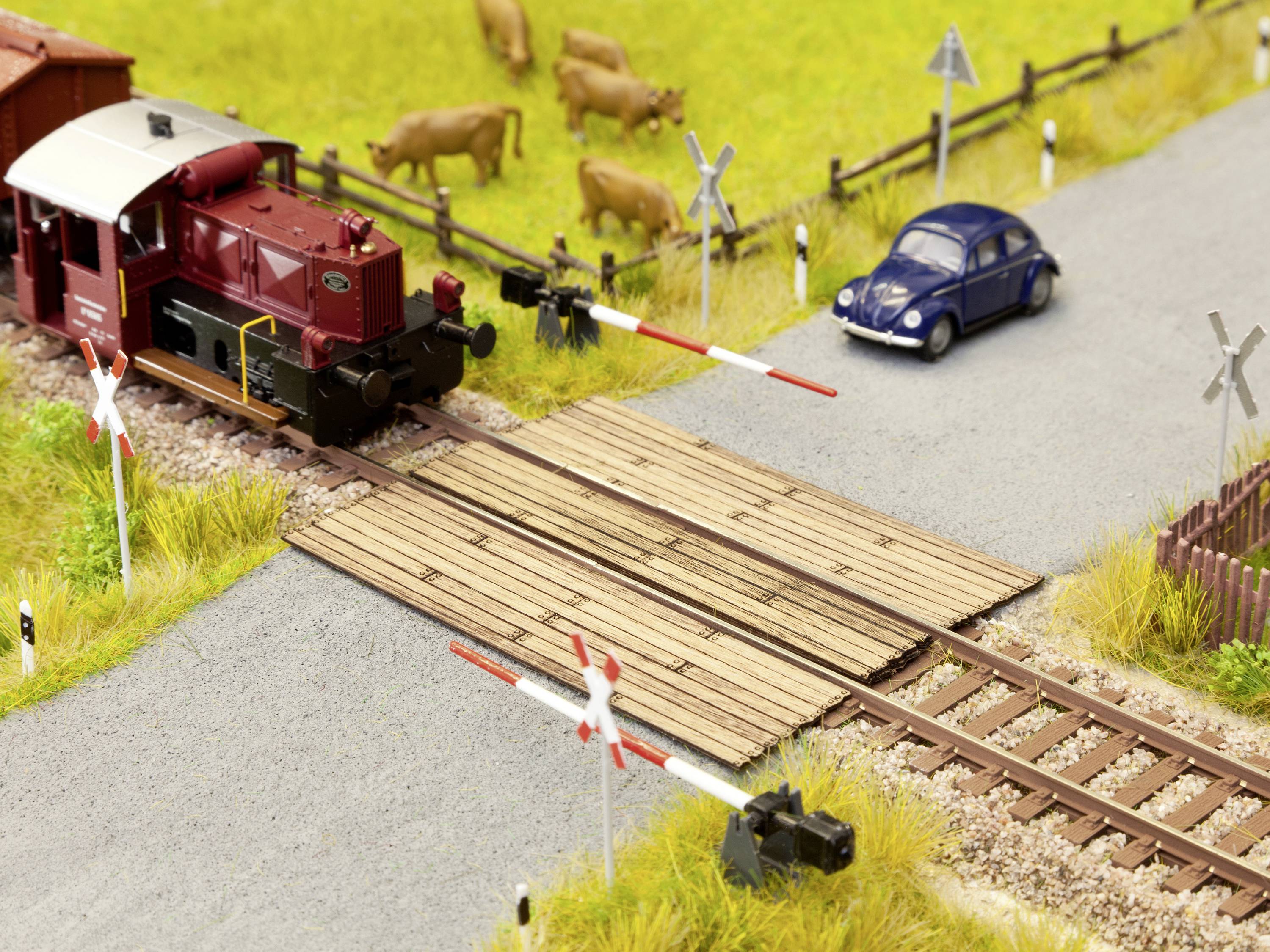Een rode trein kruist een spoorwegovergang op een modelspoorbaan. Een blauwe VW Kever wacht. Op de achtergrond grazen koeien.