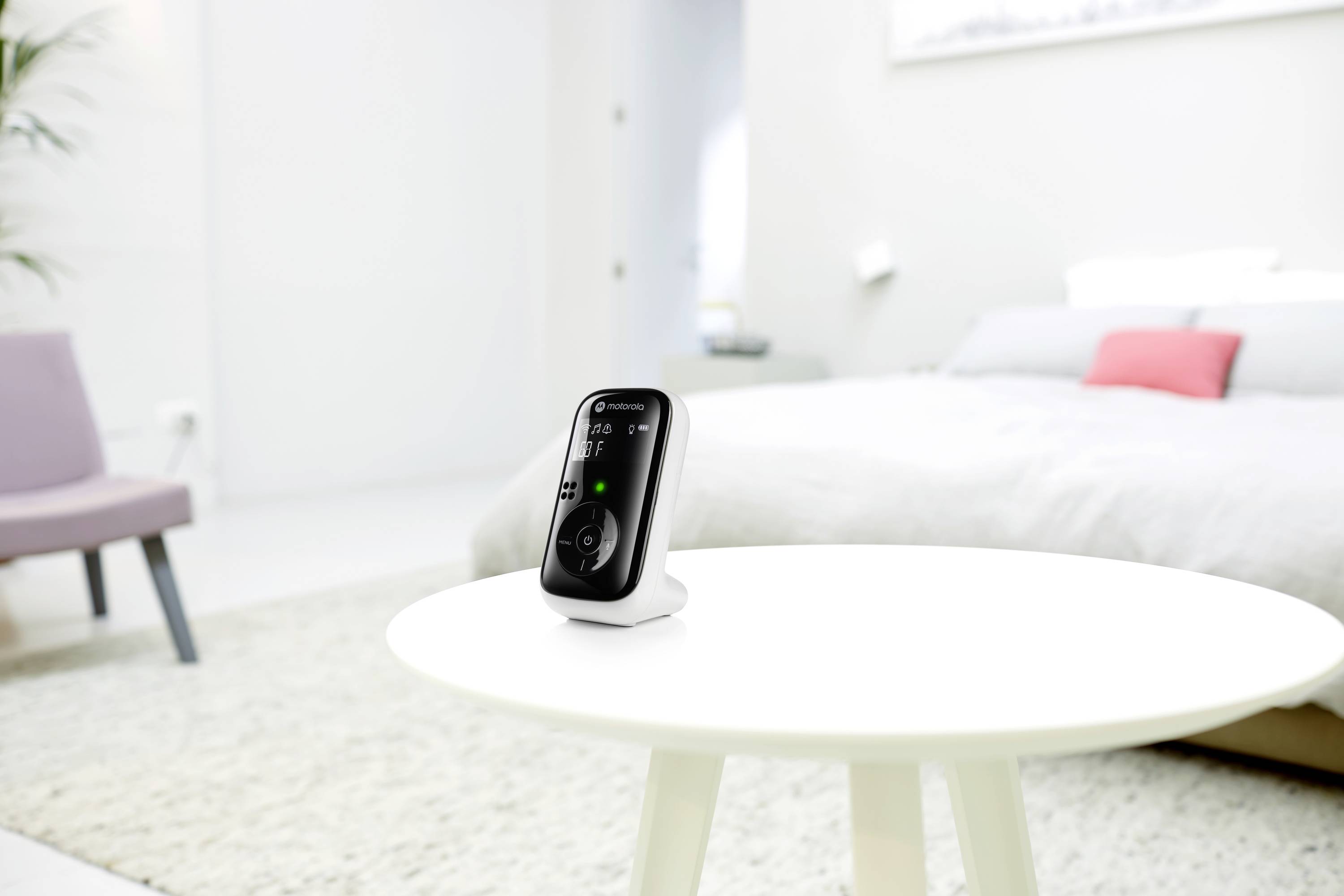 Een babyfoon staat ingeschakeld op een witte tafel in een lichte slaapkamer. Op de achtergrond is een bed zichtbaar.