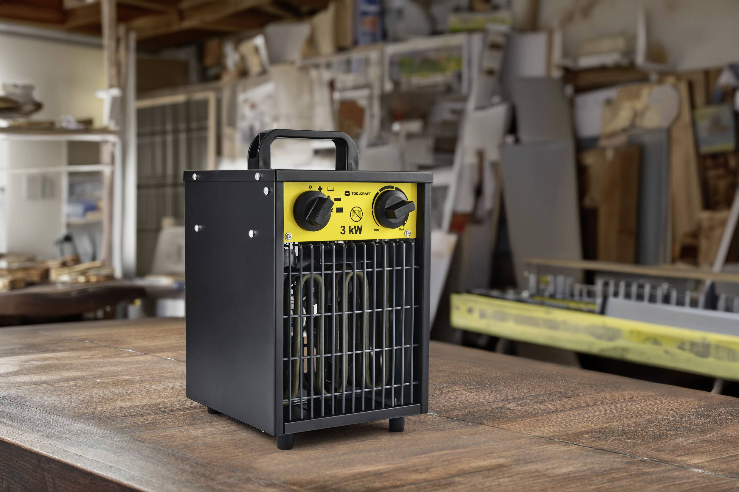 Elektrisch verwarmingstoestel van 3 kW op een houten tafel in een werkplaatsomgeving. Op de achtergrond zijn gereedschappen en materialen zichtbaar.