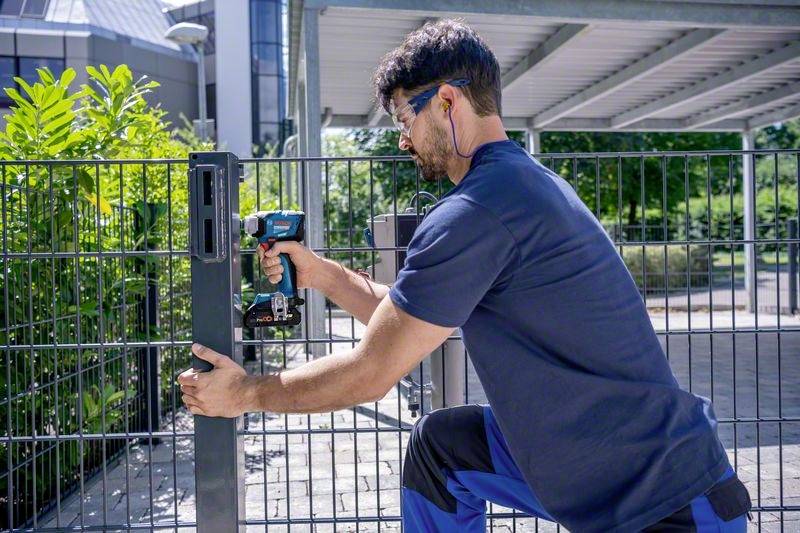 Een man in werkkleding bevestigt een metalen hek met een elektrisch gereedschap. Op de achtergrond zijn moderne gebouwen en planten zichtbaar.