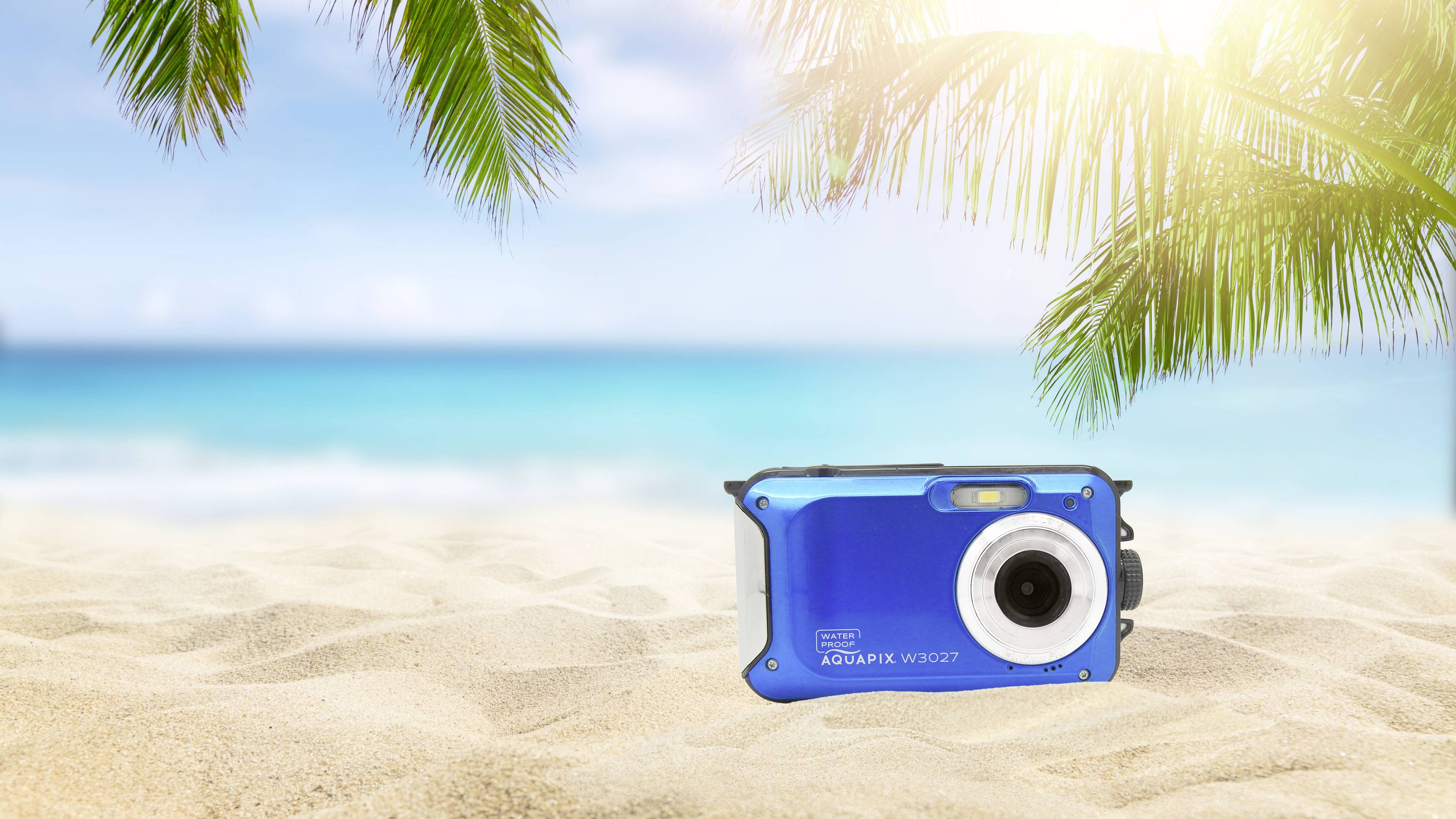 Blauwe camera ligt op een zonnig zandstrand met palmbomen en een blauwe zee op de achtergrond.