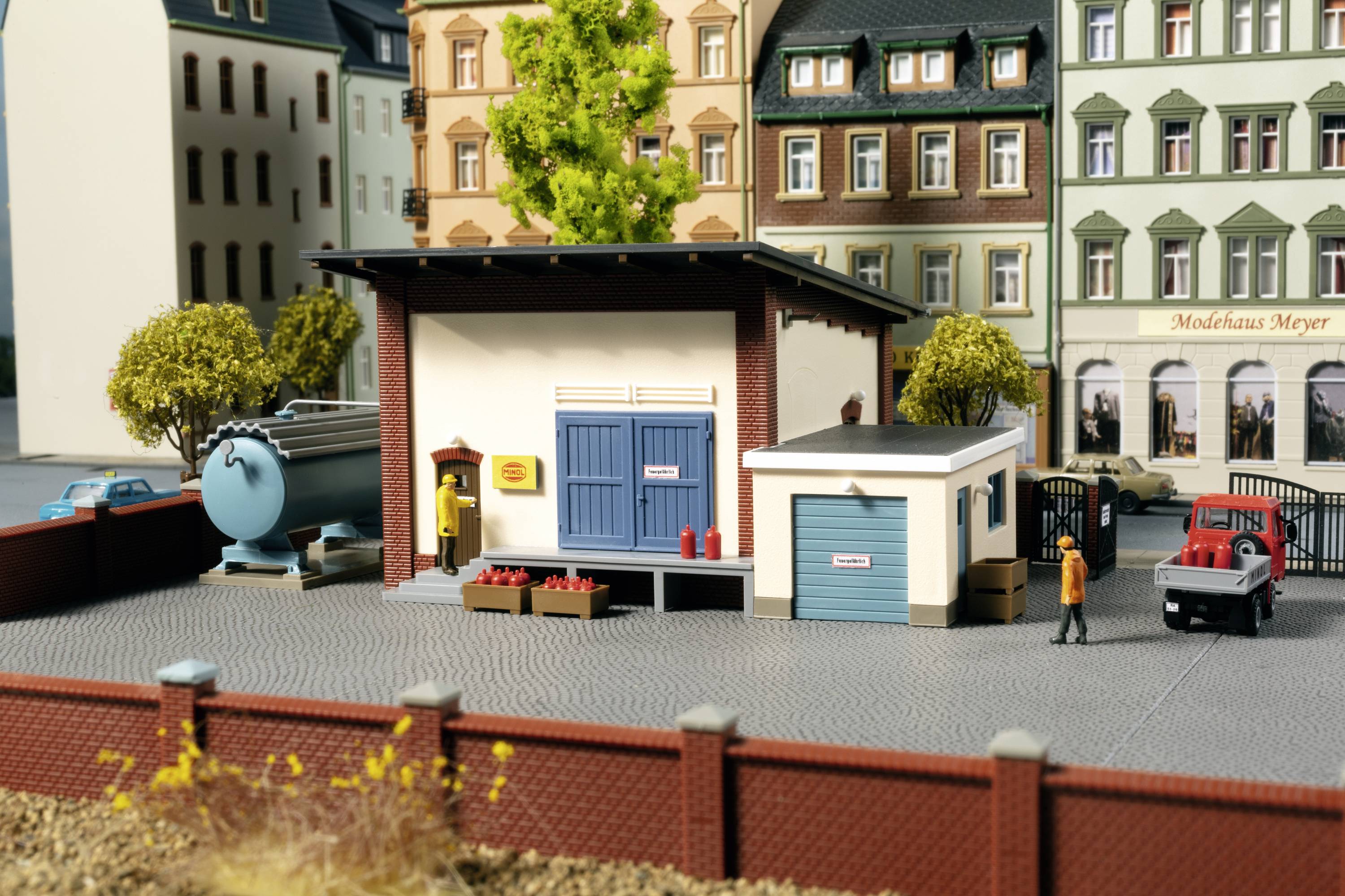 Een miniatuurscène toont een klein pakhuis op een bestrate plaats met modelvoertuigen en aangrenzende gebouwen. Twee personen staan erbuiten.